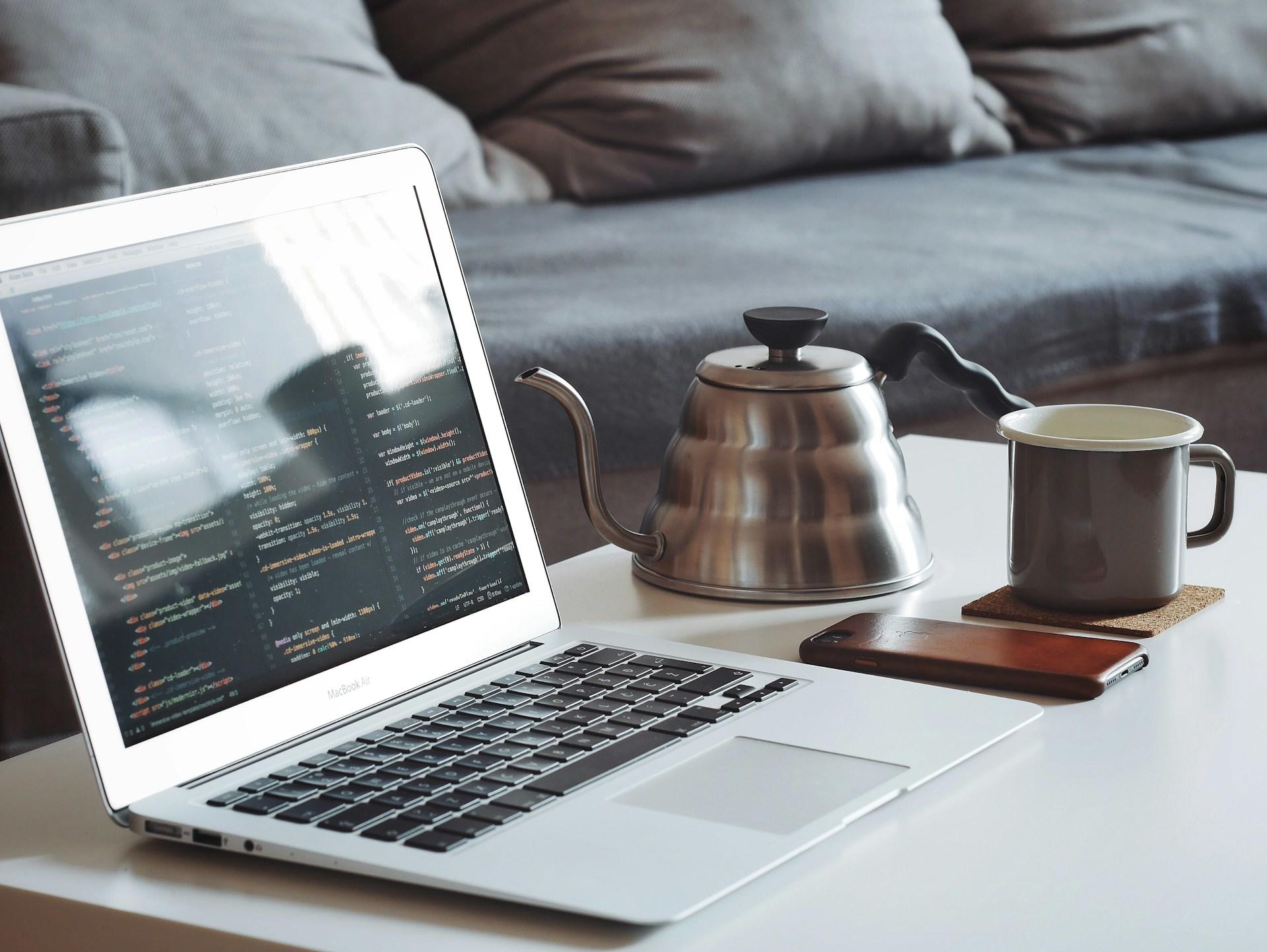 Computador em uma mesa com uma chaleira e uma xícara de café.