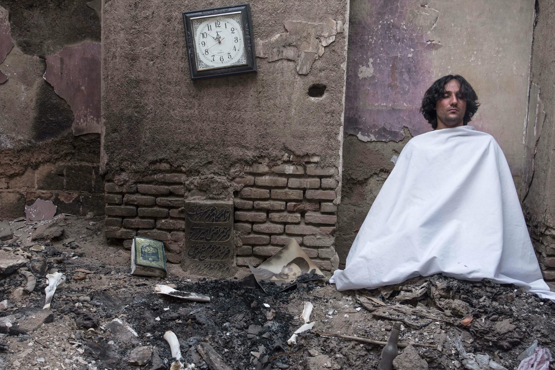 Imagem de um homem coberto por um lençol branco em um cômodo parcialmente destruído