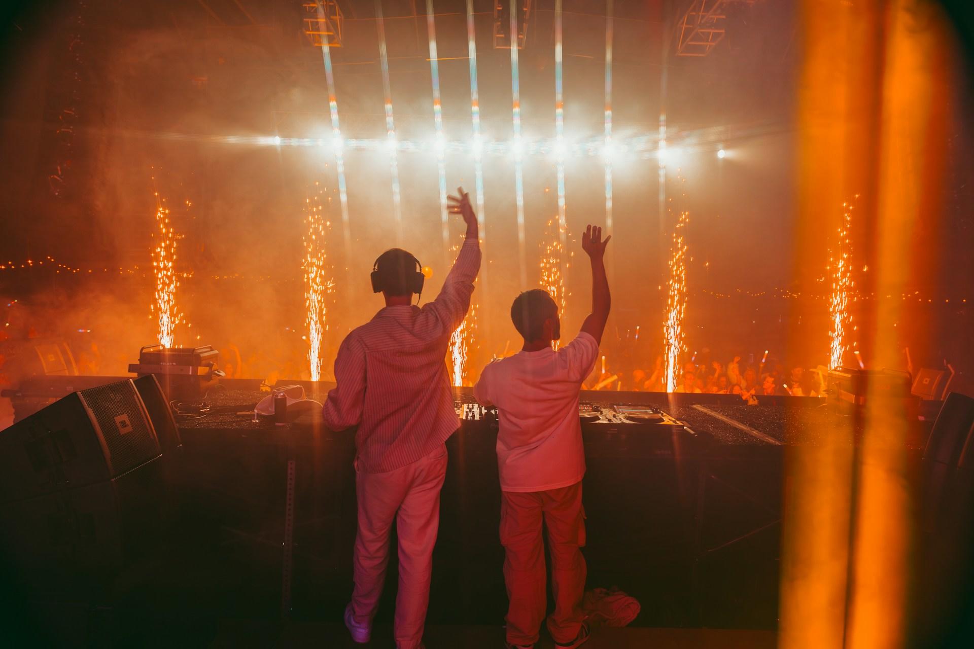 Imagem de uma festa de música eletrônica e de dois DJs comandando uma pickup.