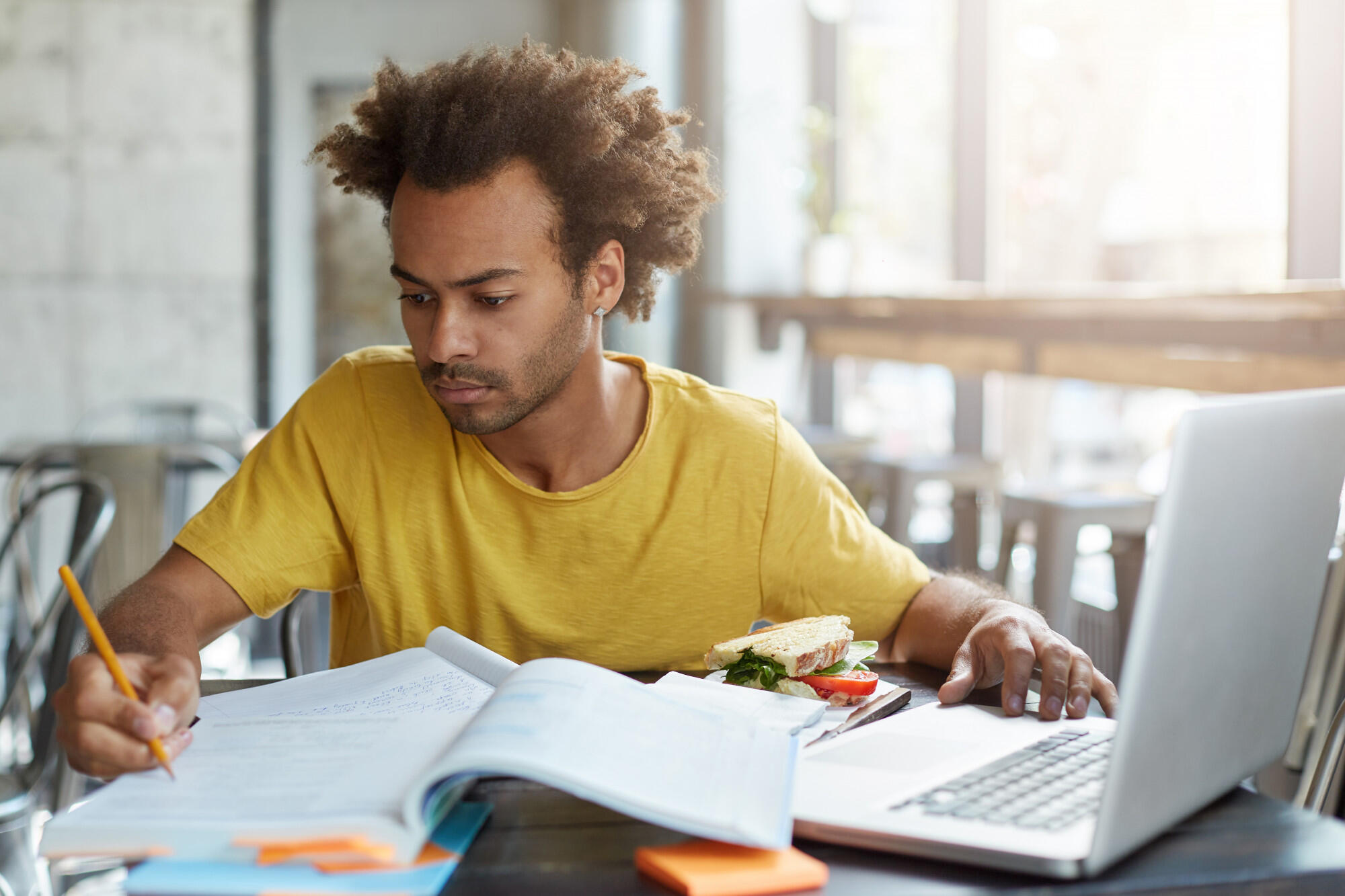 Estudante sentado estudando com o apoio do notebook e de livros. 