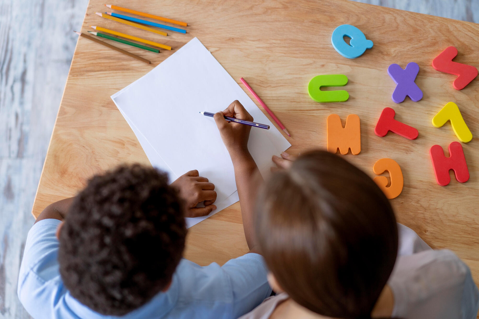 Crianças escrevendo no papel com lápis, e as letras do alfabeto ao lado.
