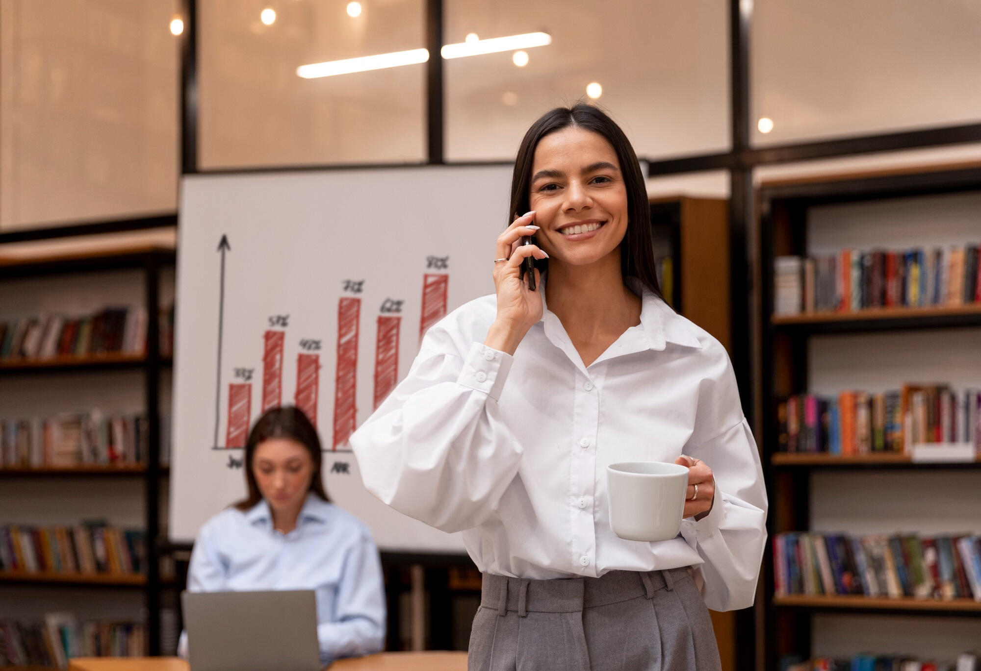 Mulher sorrindo, segurando café e falando ao telefone na sala de escritório.