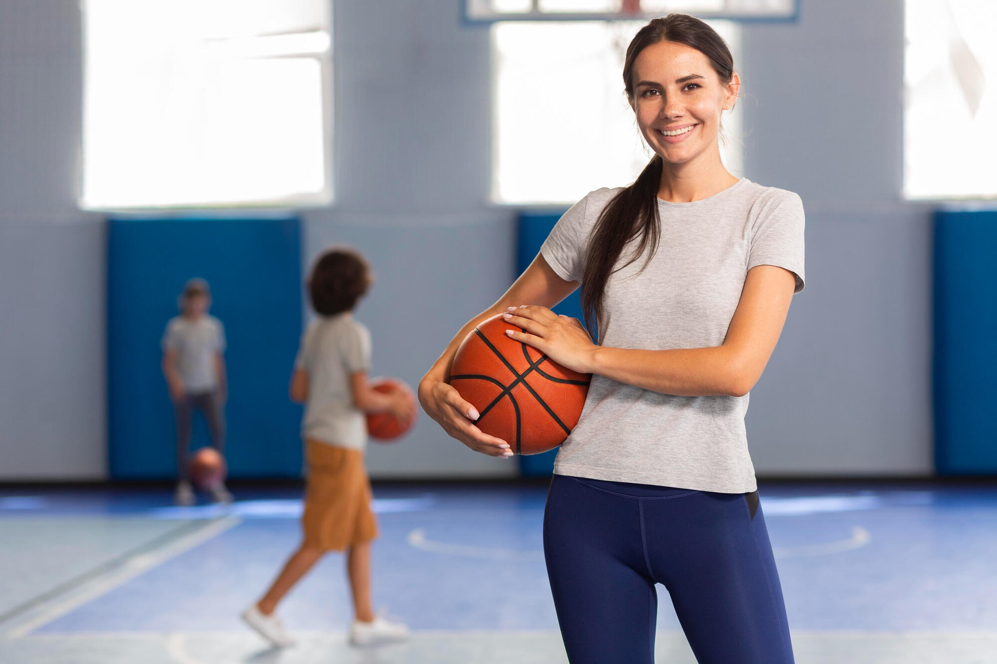 Professora de educação física com bola de basquete na mão e alunos ao fundo, na quadra.