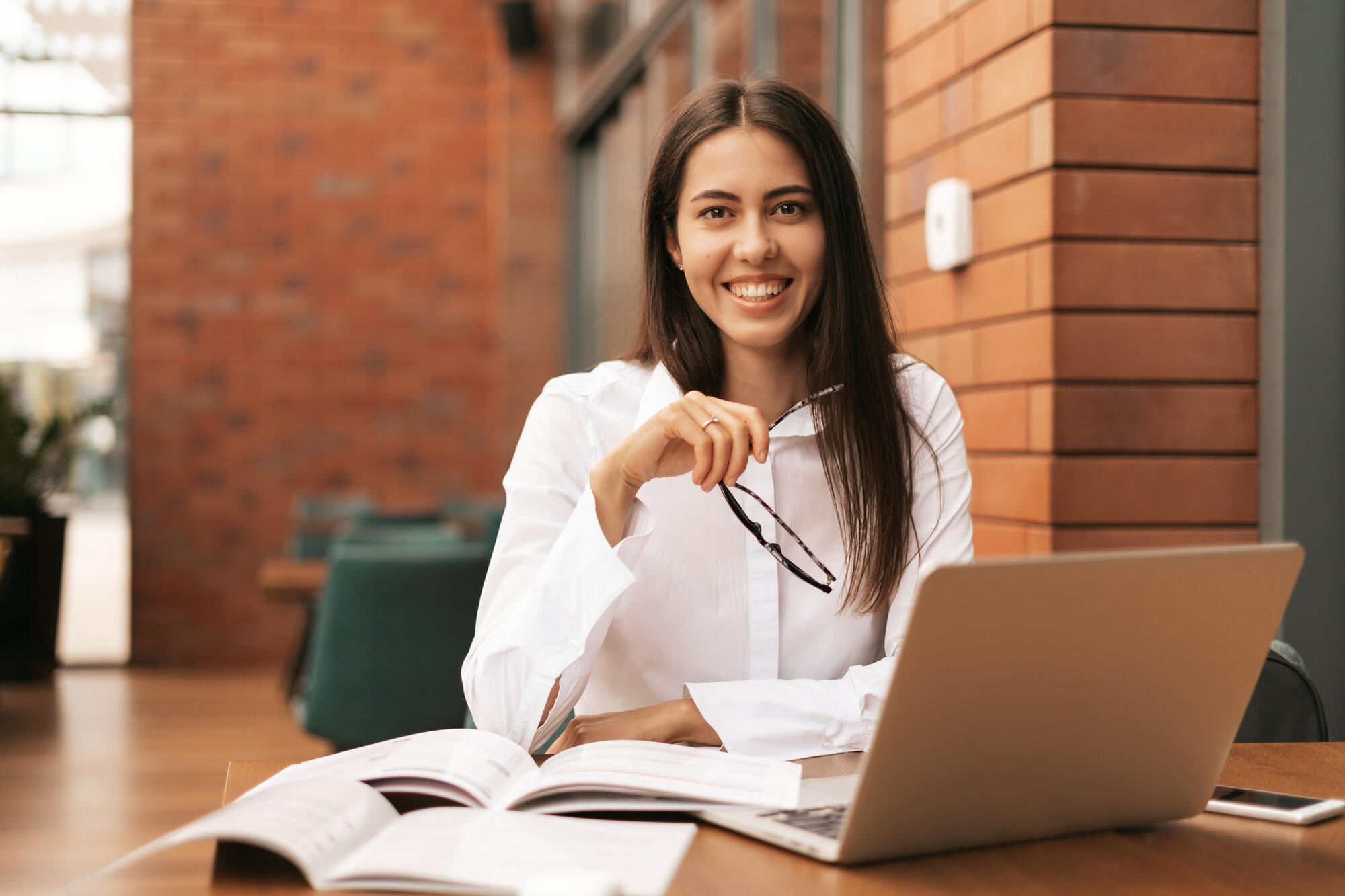 Estudante sorrindo e segurando o óculos com a mão, sentada na cadeira e com notebook sobre a mesa.
