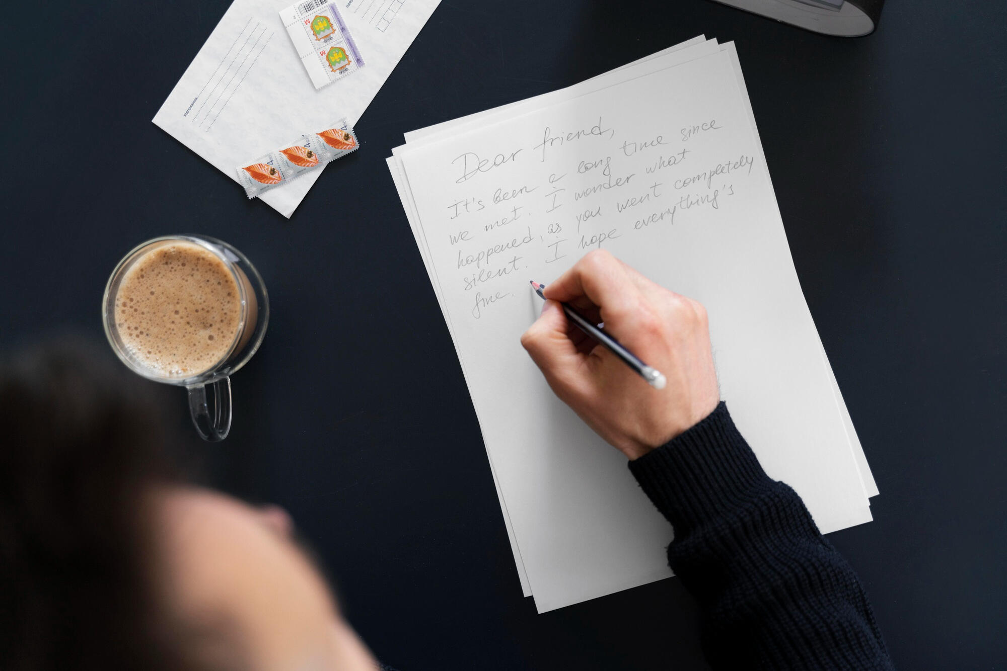 Mão de uma pessoa escrevendo uma carta, com lápis, em cima da mesa, com xícara de café ao lado.