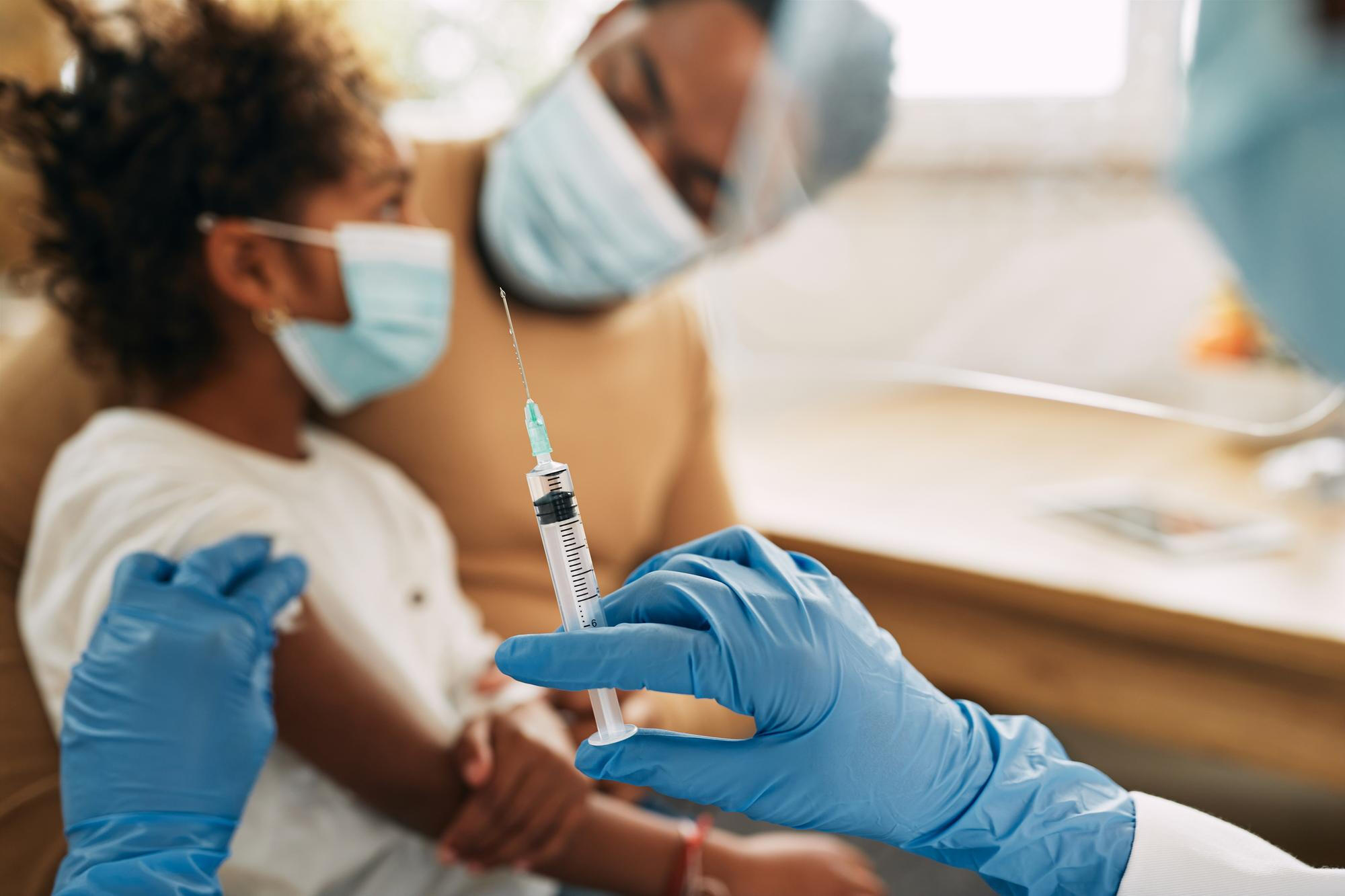 Criança com máscara prestes a ser vacinada por um profissional com luvas e seringa.