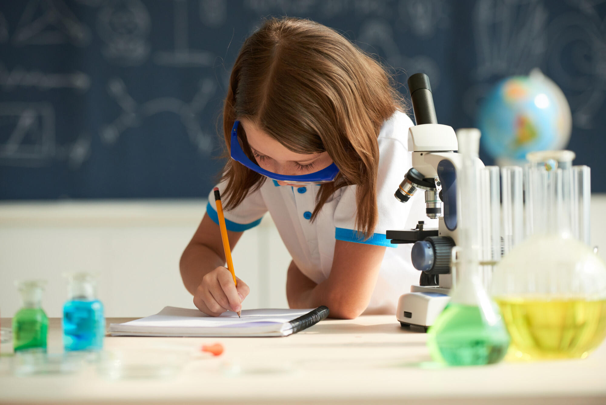 Criança estudando ciências, escrevendo no papel, na mesa com miscroscópio ao lado e lousa ao fundo.
