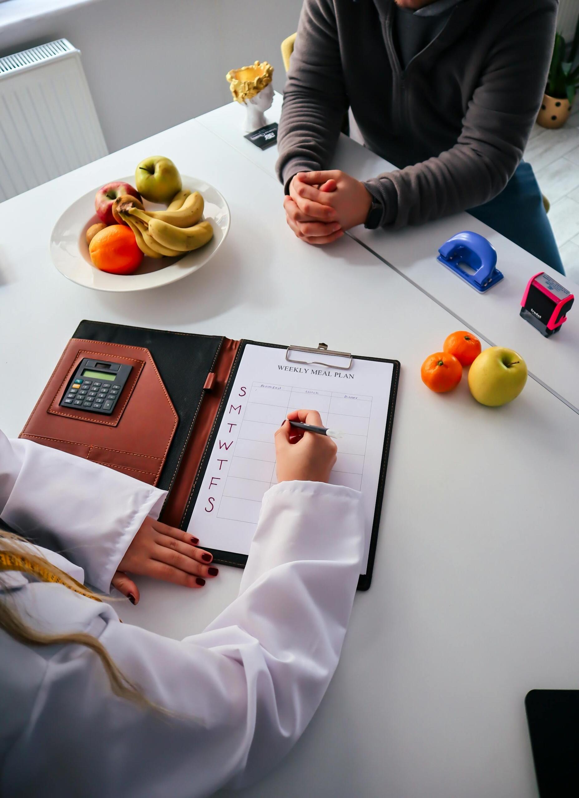 A person's hand writes on a weekly meal plan while a plate of fruits sits on the table, and another person listens attentively.