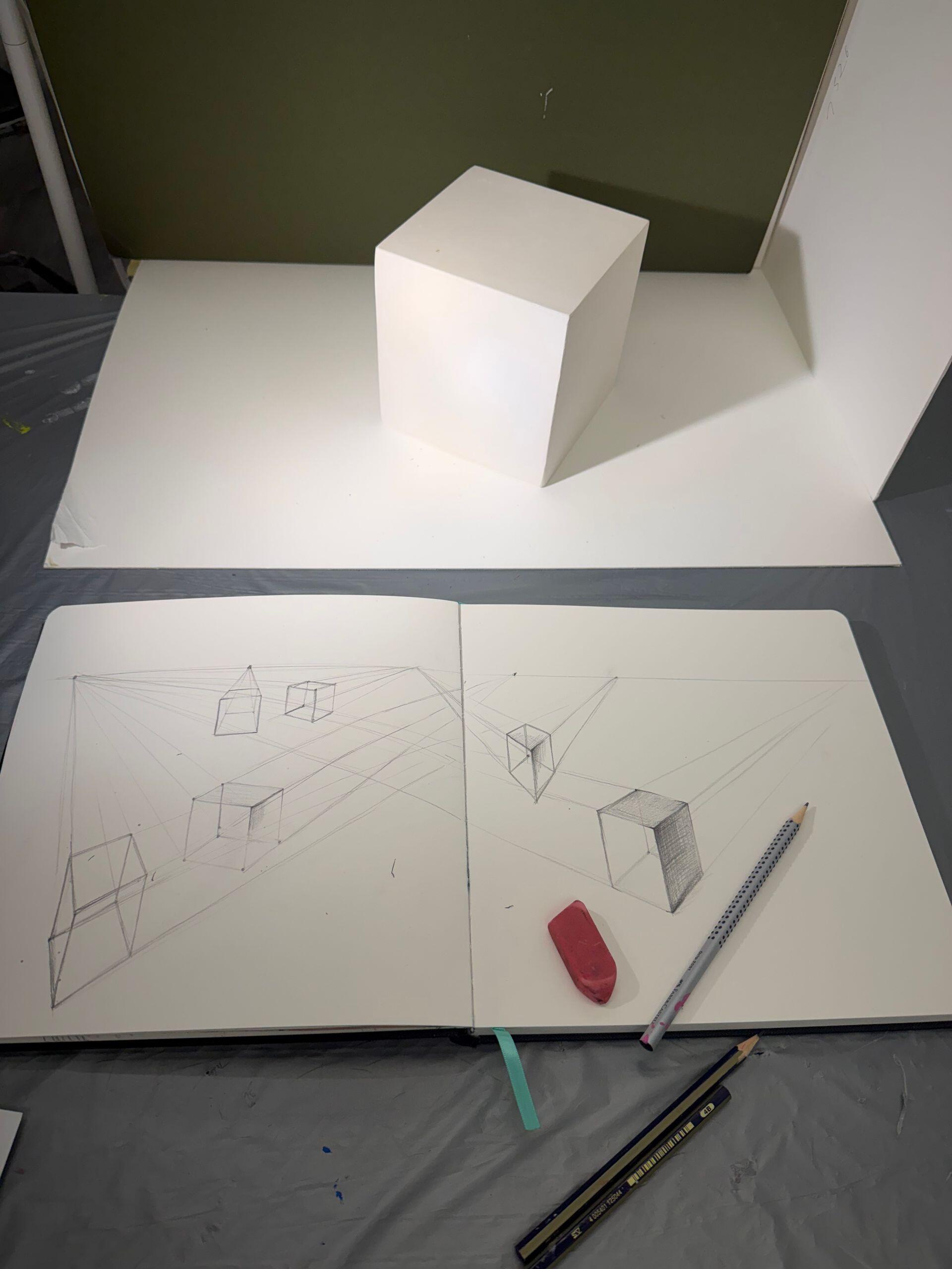 A white cube is positioned on a white surface with an open sketchbook in the foreground showing perspective line drawings of cubes, accompanied by a pink eraser and pencils.
