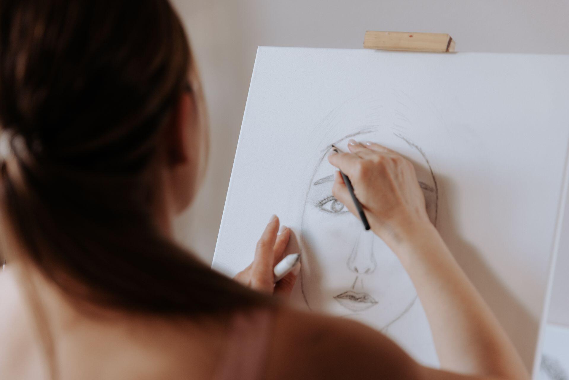 A woman with dark hair stands before an easel, using a pencil and blending tool to sketch a portrait on a canvas.