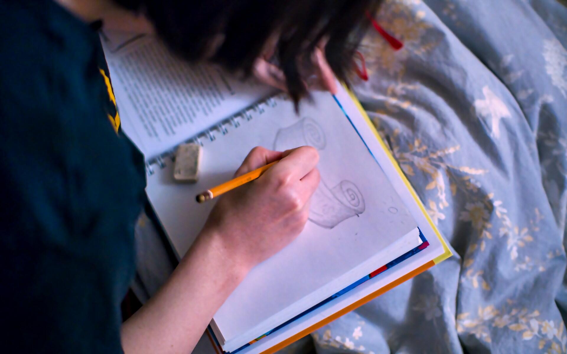 High-angle shot of a person sketching a scroll with an orange pencil in a notebook while sitting on a patterned bed.