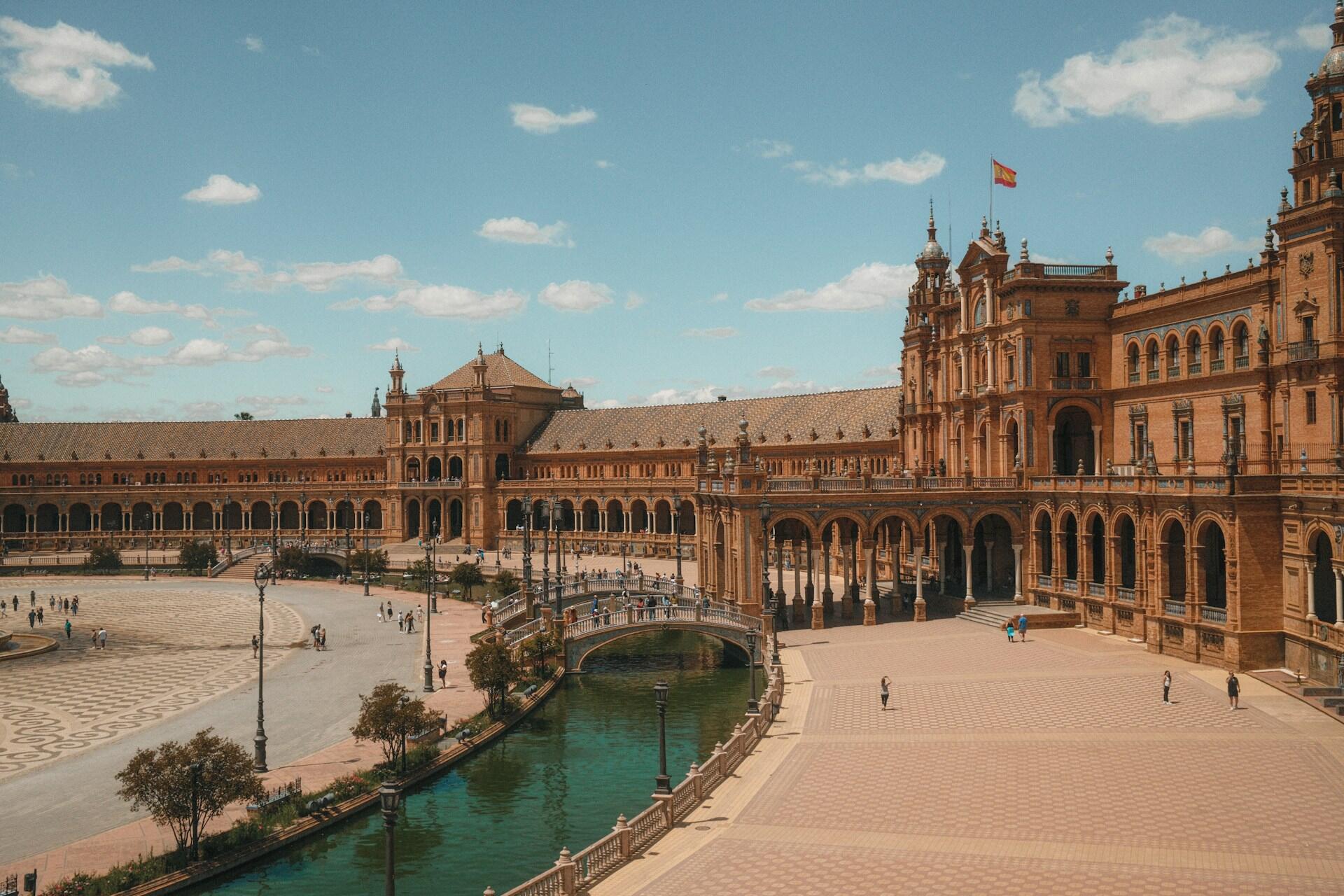 Plaza de España in Seville, Spain, showcasing its large semi-circular building, canal, bridges, and people under a partly cloudy sky.