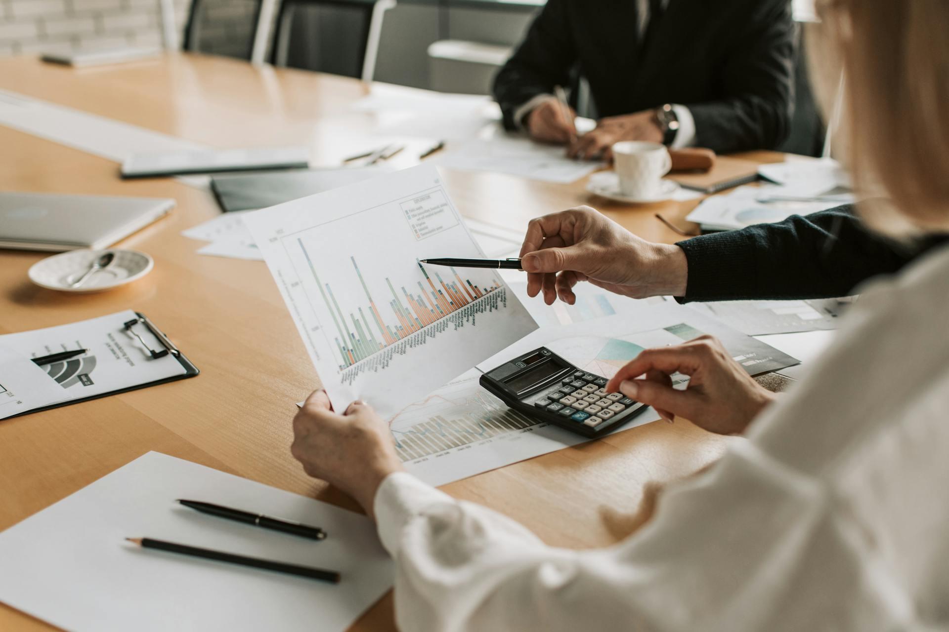 Close-up of business professionals reviewing a bar chart document with a calculator on a light wooden conference table.