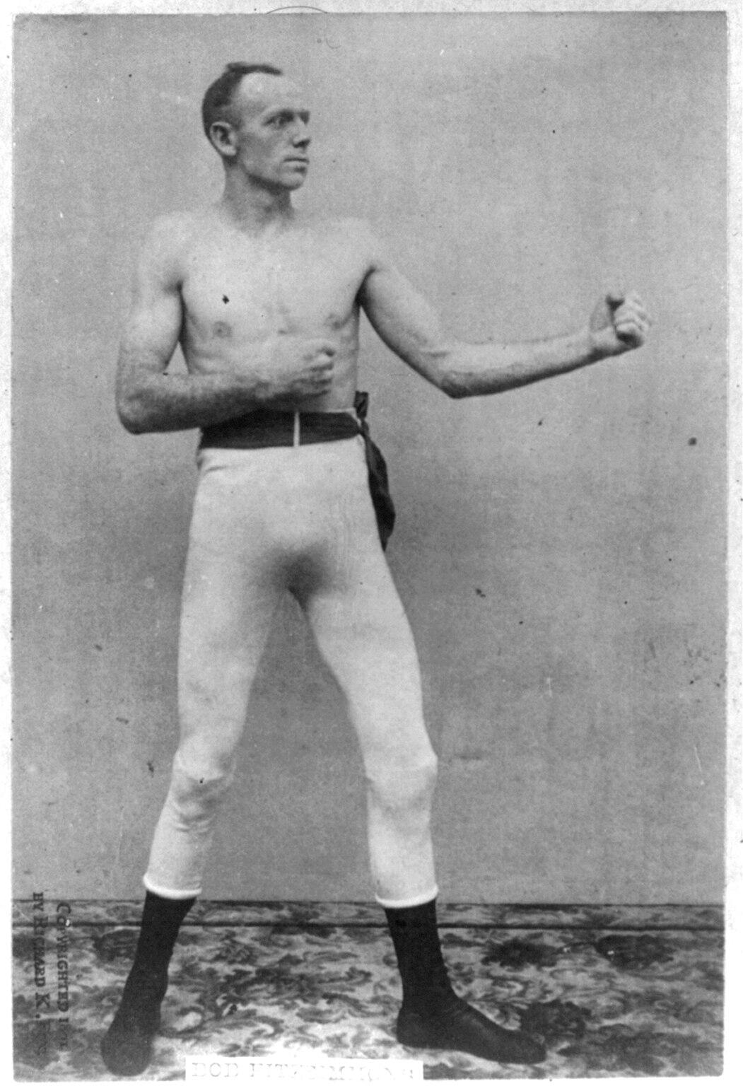A muscular man poses in a vintage-style boxing stance, wearing white pants and black socks, with a plain background.