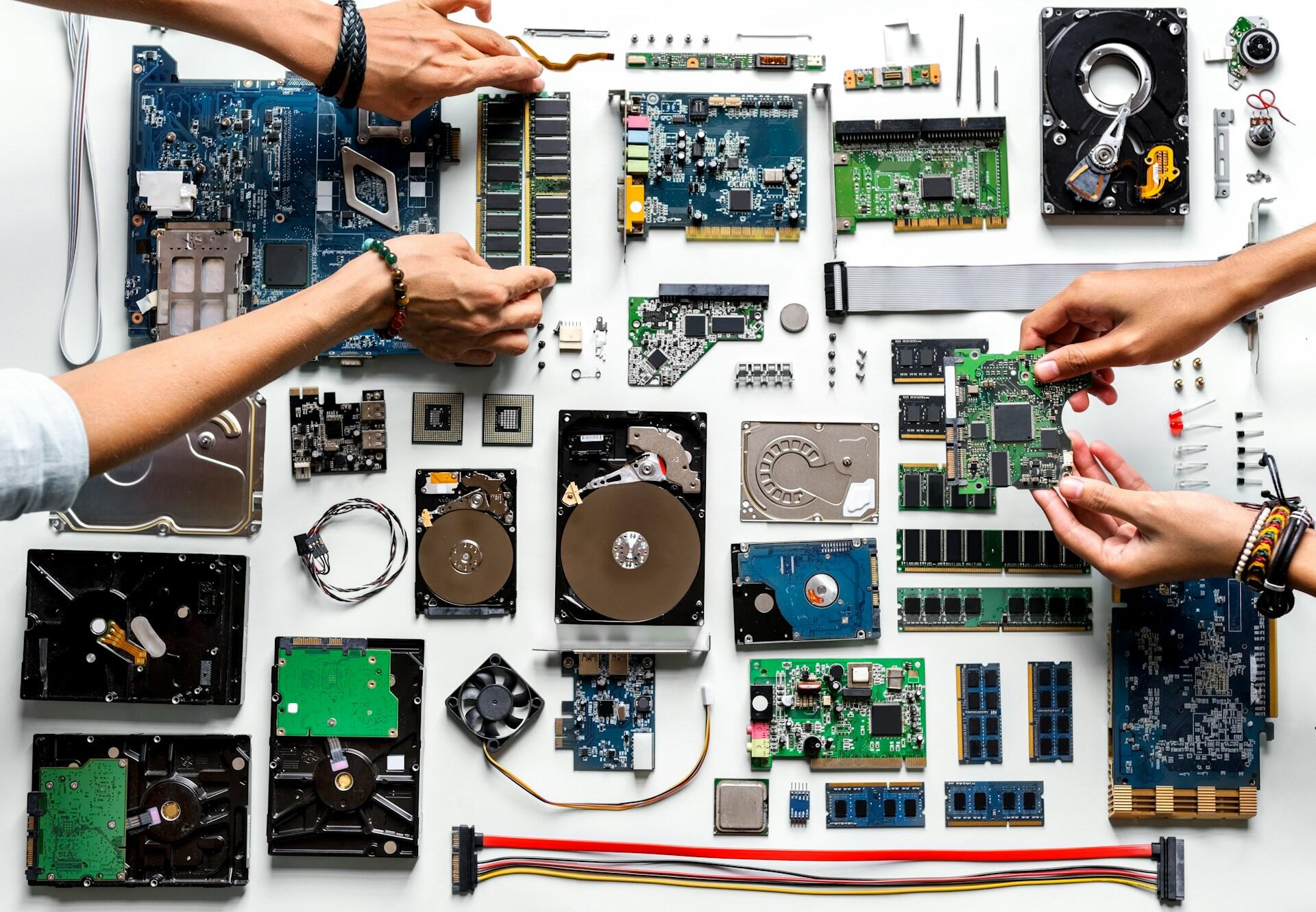 A flatlay of computer components including circuit boards, hard drives, and memory modules, with hands assembling parts.