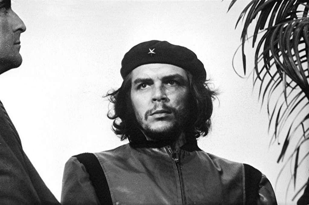 Che Guevara in a military-style jacket and cap stands beside another individual, with a plant visible in the background.