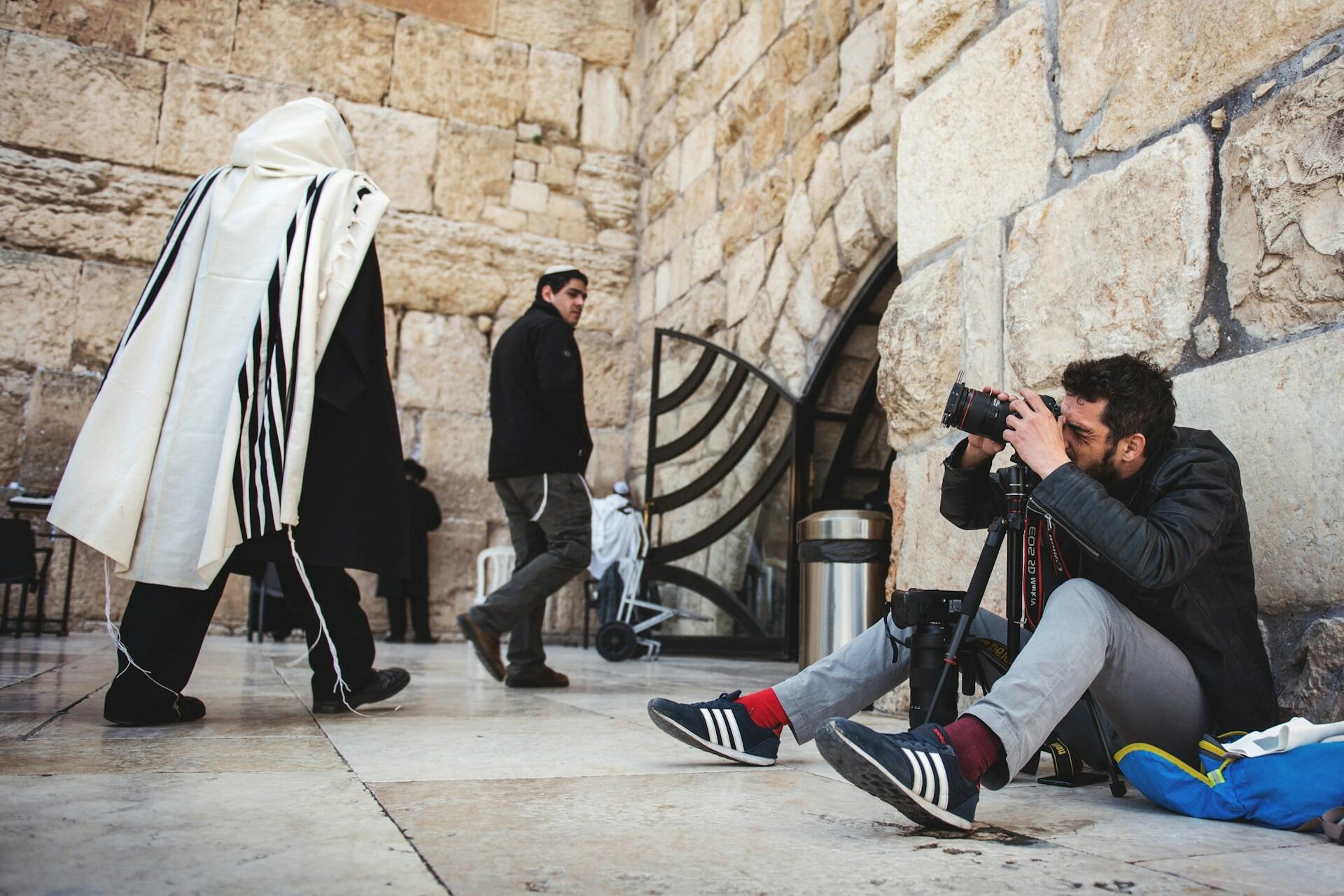 A photographer crouches against a stone wall, capturing images at a busy historic site while people walk by.
