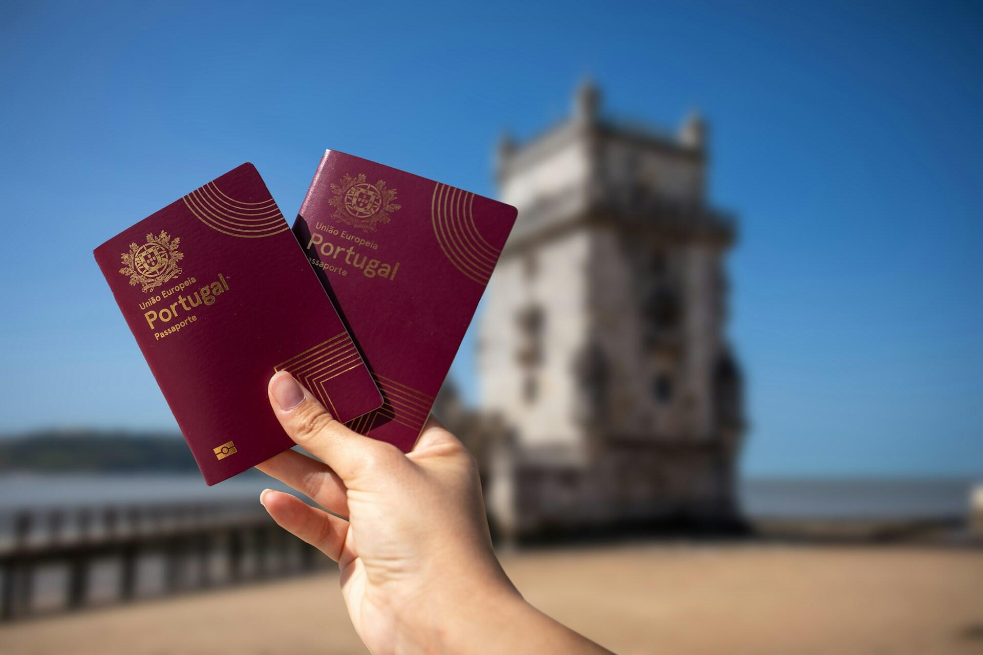 Hand holding two maroon Portuguese passports with a historic tower and clear blue sky in the background.
