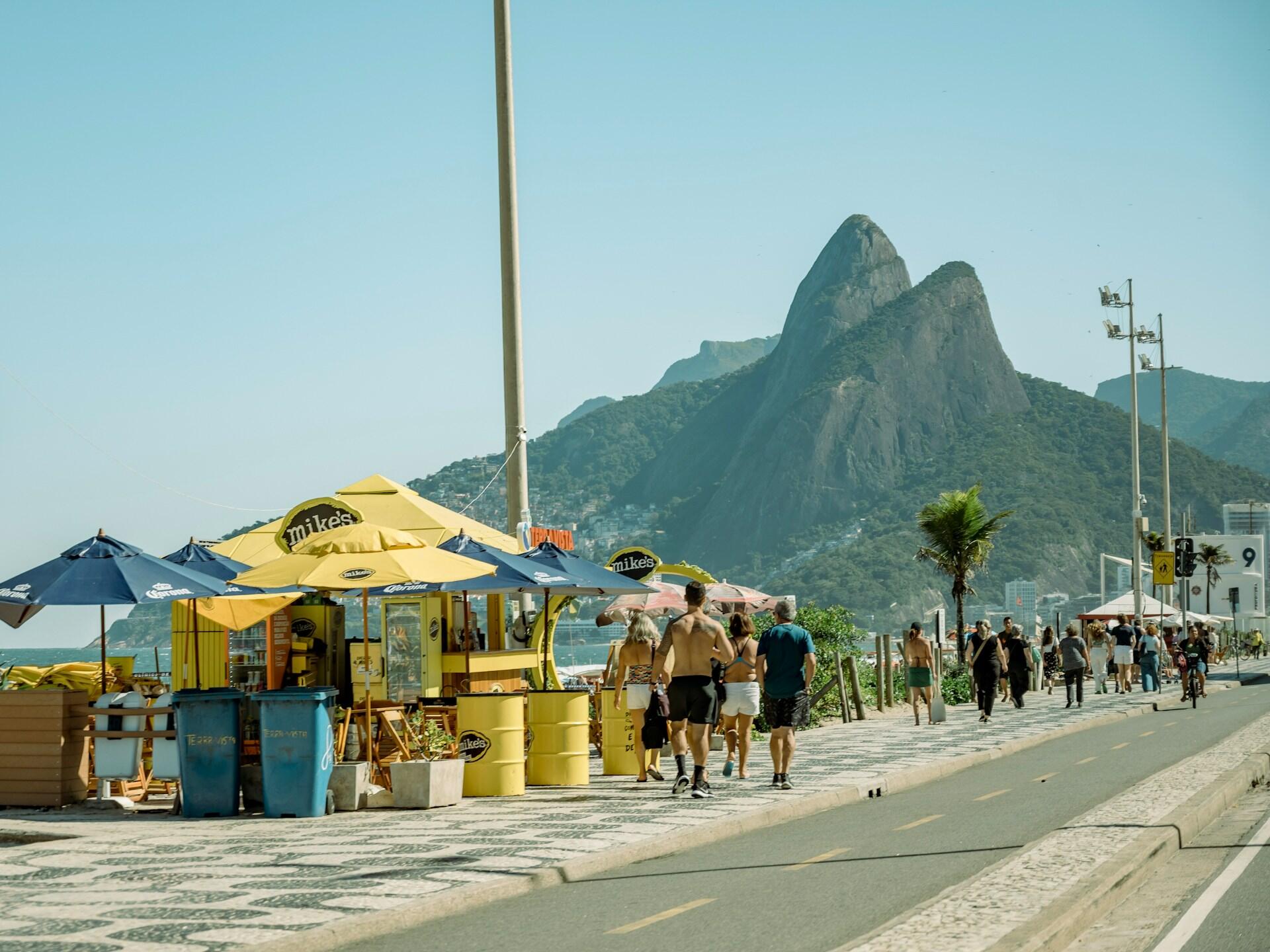People stroll along a beachside street with mountains rising in the background, enjoying a sunny day outdoors.