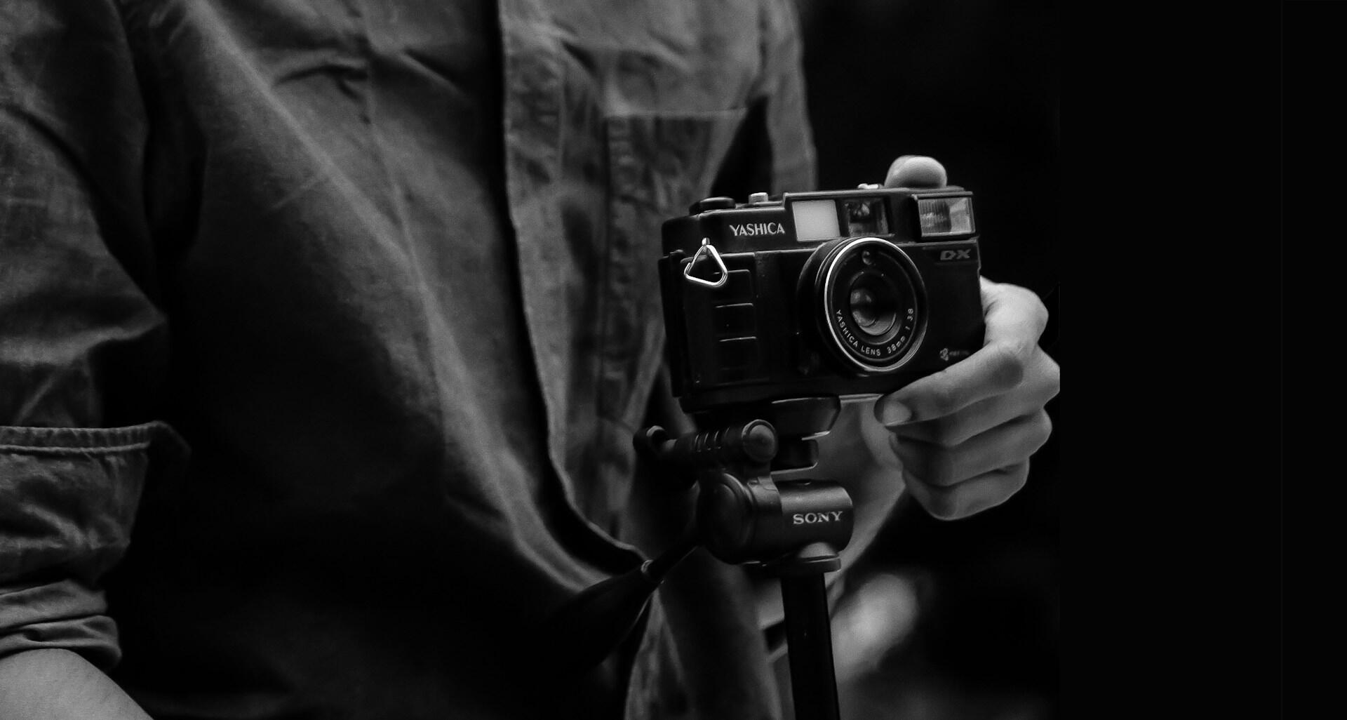 A hand holds a vintage Yashica DX camera mounted on a tripod, set against a dark background, evoking a nostalgic feel.