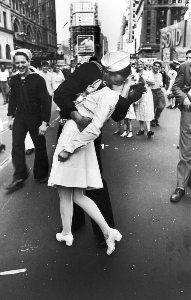 A sailor embraces a woman in a white dress, passionately kissing in a lively, historical New York street scene.