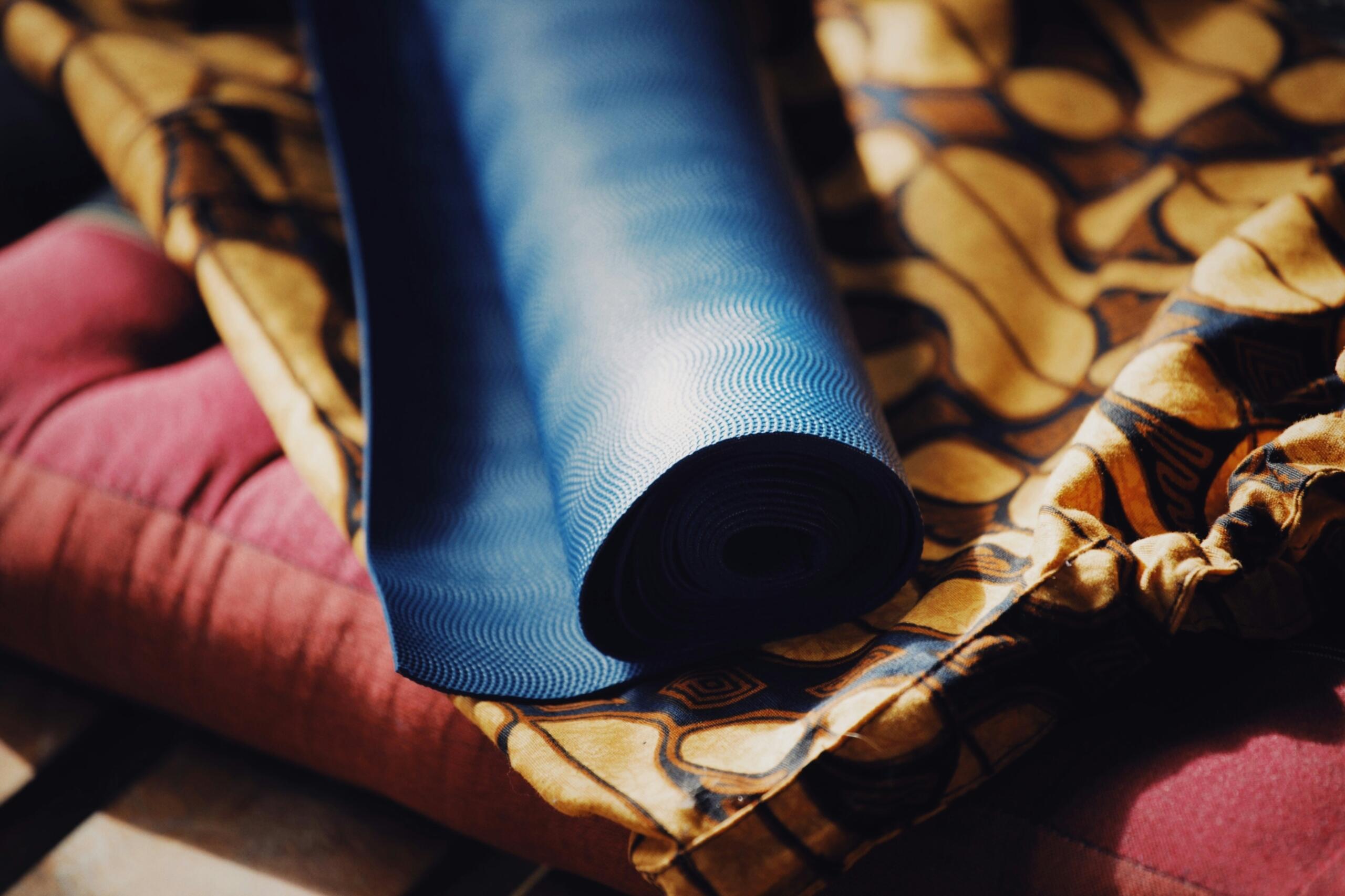 A blue yoga mat is rolled up alongside vibrant patterned fabrics, resting on a cushioned surface with warm tones.