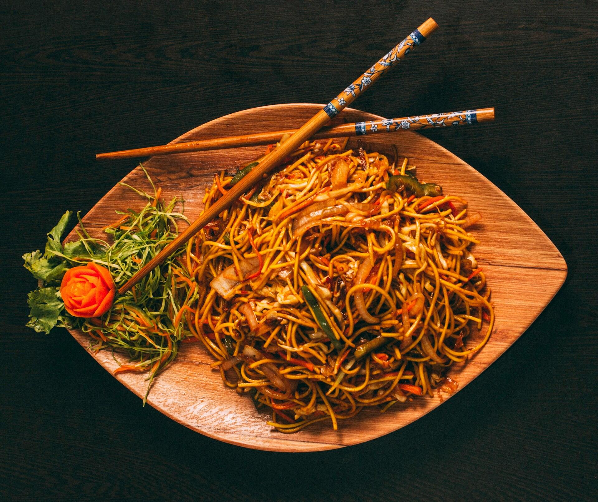 A wooden platter overflowing with stir-fried noodles, garnished with vibrant vegetables and a rose-shaped carrot, alongside decorative chopsticks.