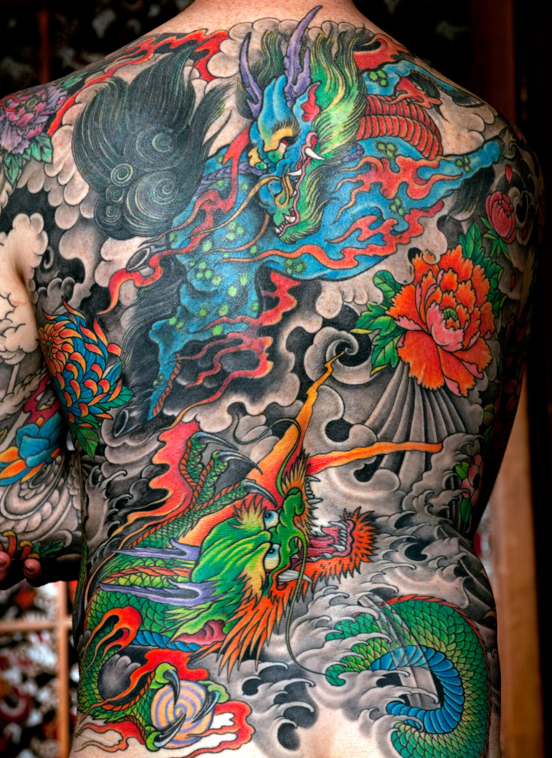 Artistic full-body Japanese irezumi tattoo on a man’s back, showing a vibrant dragon and phoenix surrounded by waves, clouds, and peony flowers.
