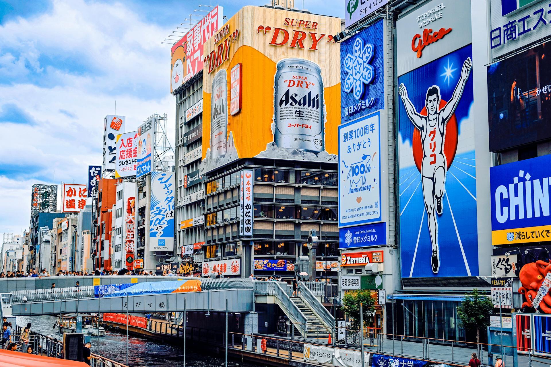 Vibrant urban canal scene in Japan with colorful billboards, including Asahi beer and Glico running man, and a pedestrian bridge over the water.