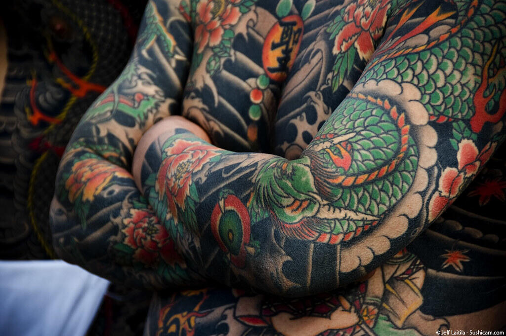 Close-up of a person's arms and torso covered in colorful traditional Japanese-style tattoos featuring flowers and a green dragon.