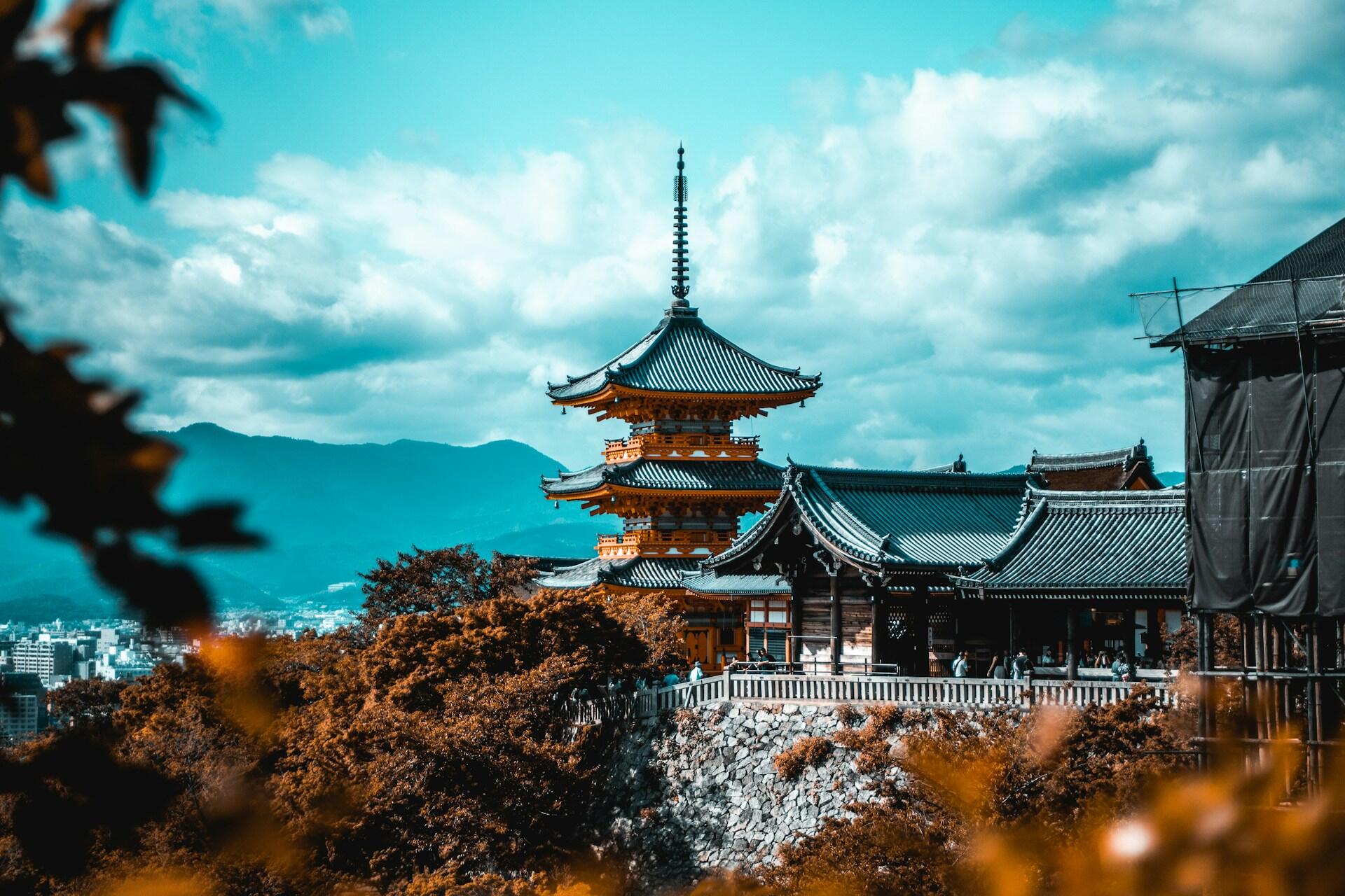 Traditional Japanese pagoda and temple complex on a stone terrace surrounded by autumn foliage with mountains under a cloudy sky.