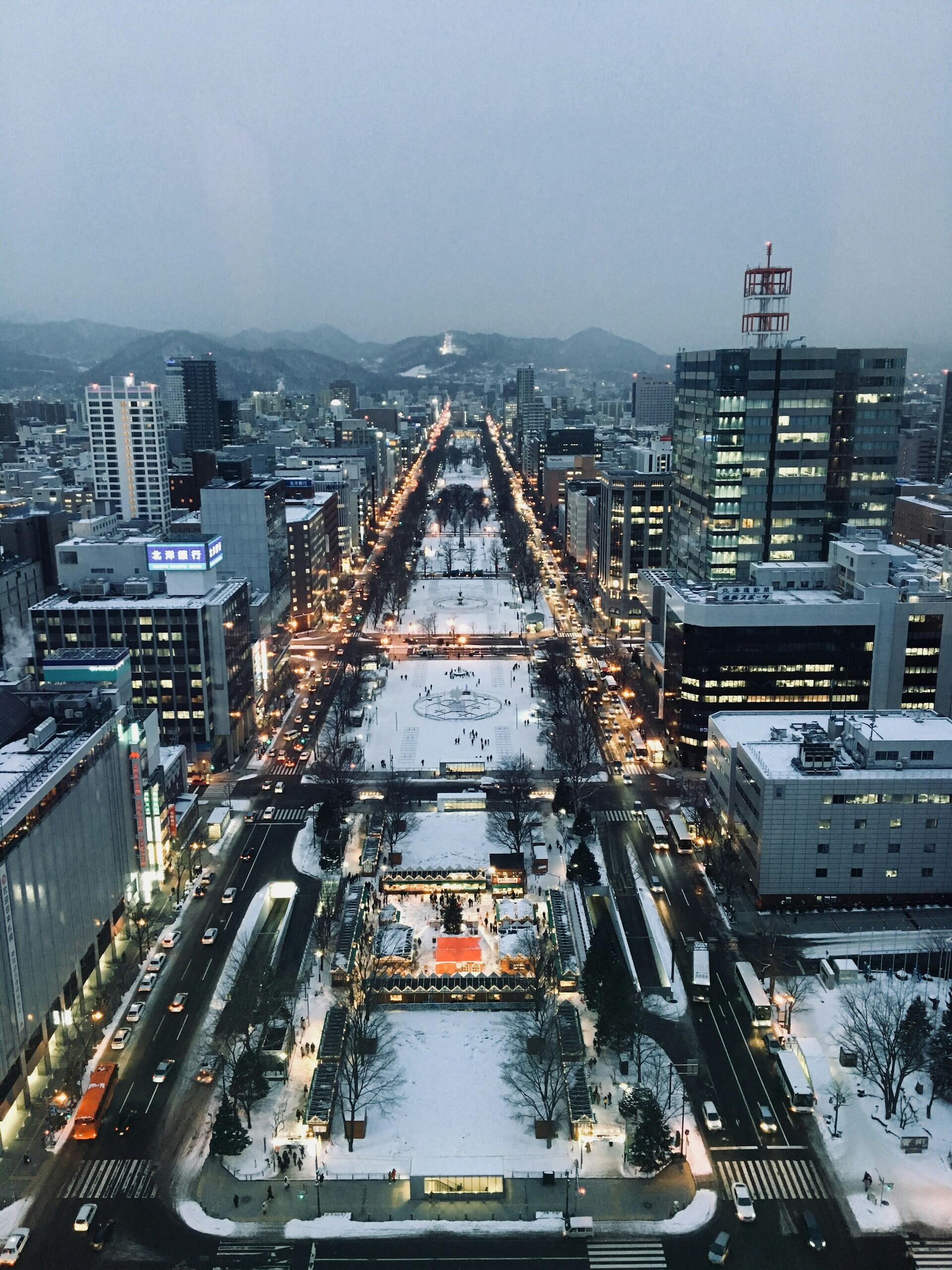Snow-covered urban park with lit pathways and surrounding high-rise buildings during dusk in a city with mountains in the background.