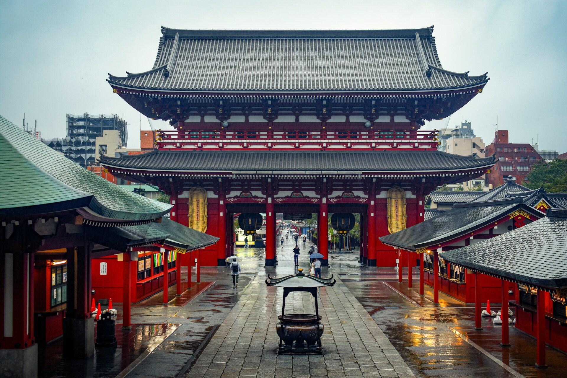 Red traditional Japanese temple gate and courtyard with people holding umbrellas on a rainy day.