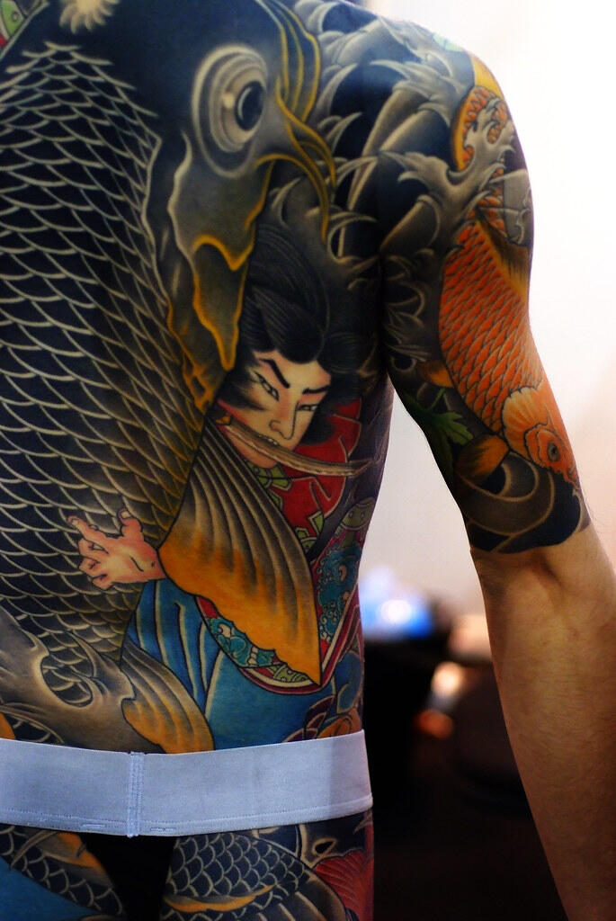 Intricate full-back and arm Japanese-style tattoo featuring a koi fish and a traditional warrior in vibrant colors.