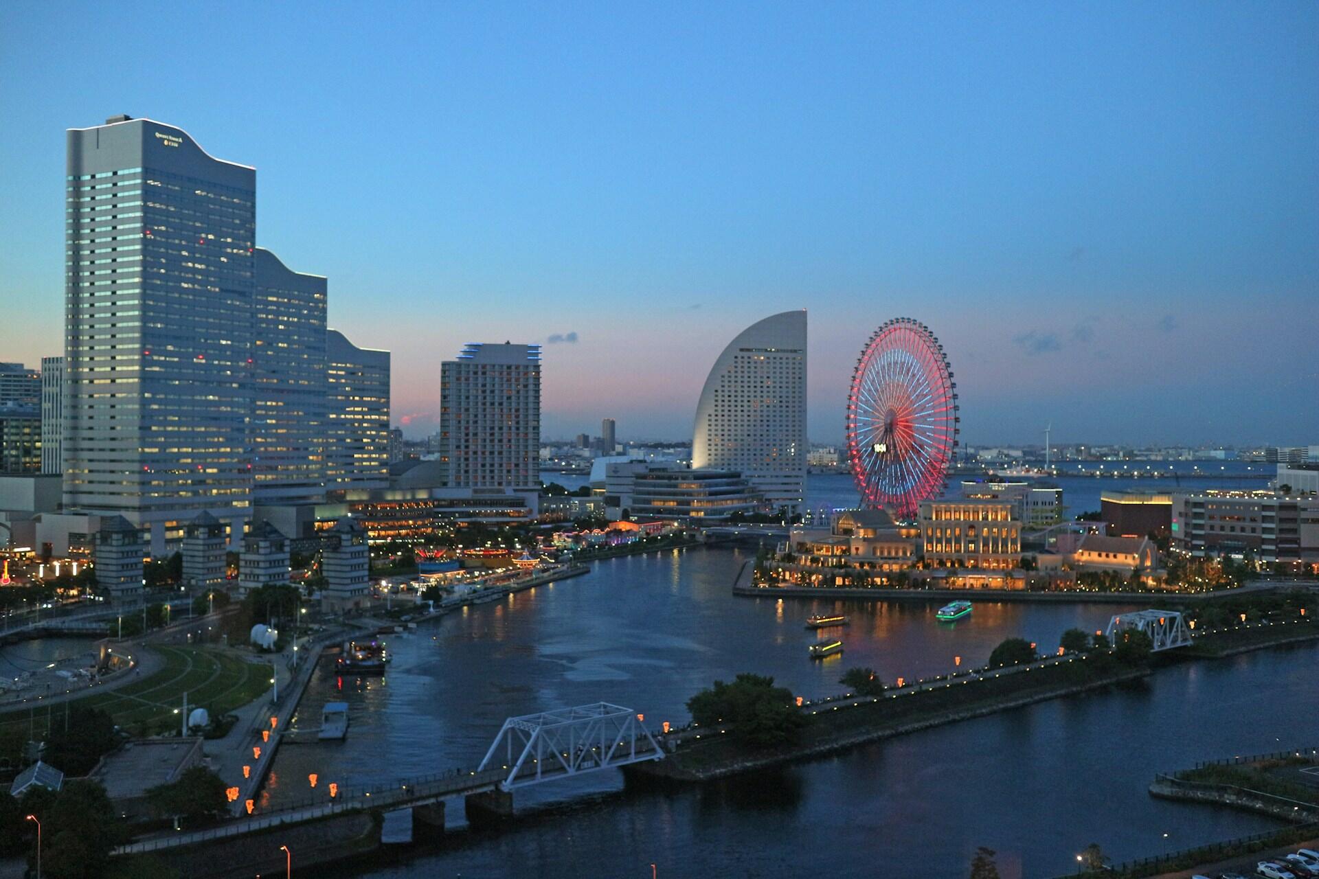 City waterfront at dusk with illuminated Ferris wheel, modern buildings, and calm river reflecting lights.