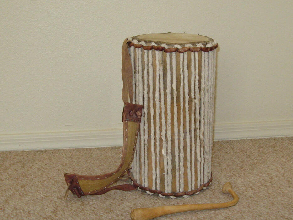 A traditional West African talking drum with tension cords and a curved playing stick placed on the floor.