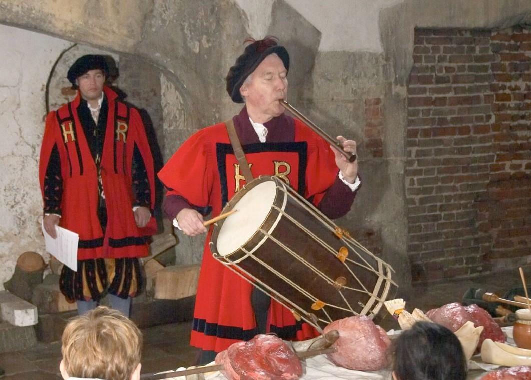 A reenactor in period costume plays a tabor drum with one hand while blowing into a pipe flute.