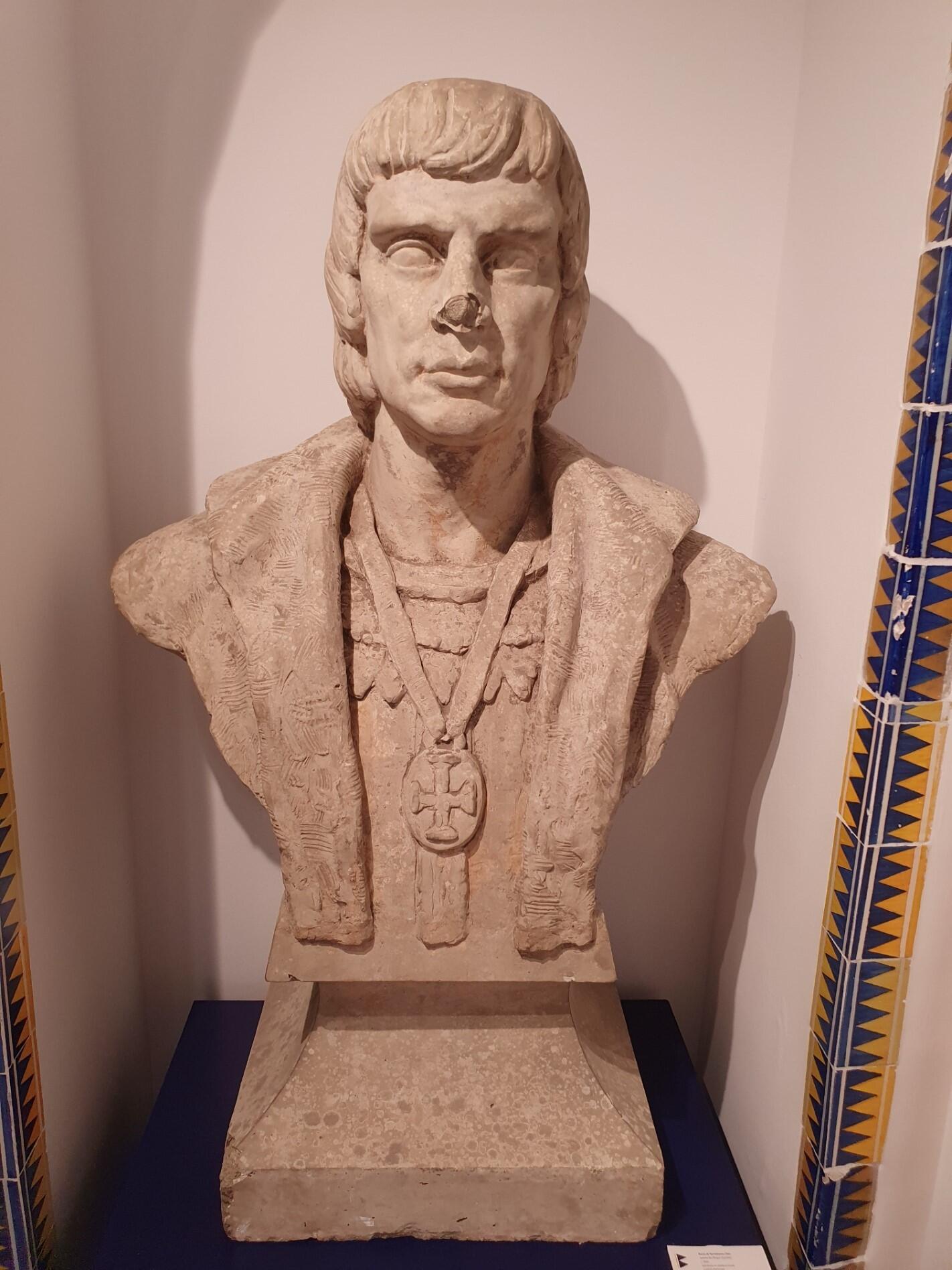 A stone bust of a man with short, straight hair, wearing a textured cloak and a medallion featuring a cross. The statue is displayed indoors on a pedestal, with patterned tiles partially visible on either side.