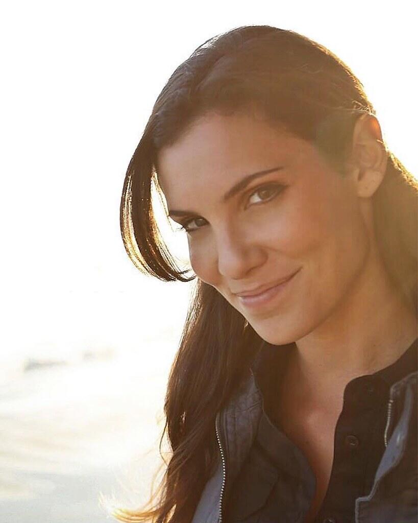A woman with long dark hair wearing a stylish vest stands outdoors, illuminated by warm sunlight against a serene background.