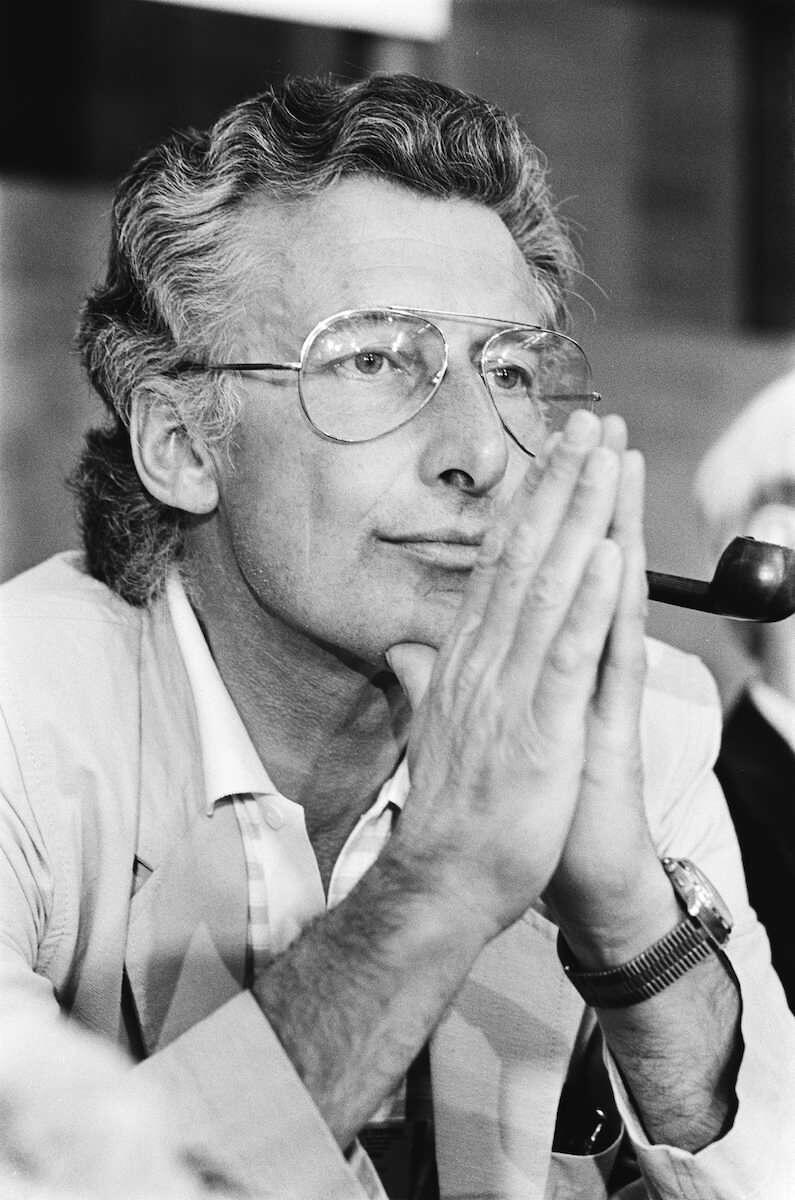 Black-and-white portrait of Dutch author Harry Mulisch wearing large aviator-style glasses and a light-colored blazer, resting his clasped hands near his face while holding a pipe. He looks thoughtfully ahead, with a blurred indoor background suggesting a public event or literary gathering.