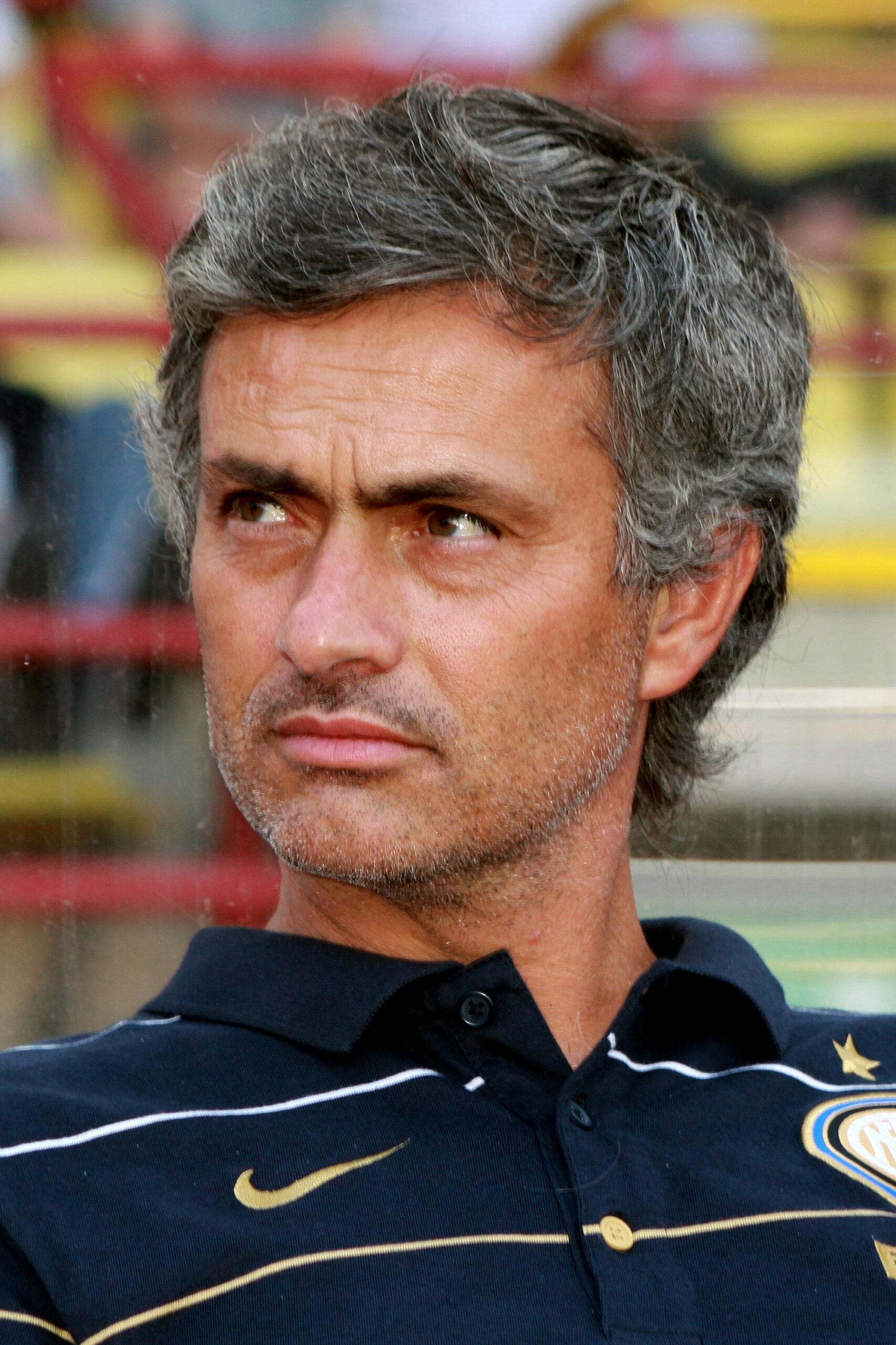 A man with short gray hair wearing a navy blue sports polo shirt with gold details and a crest, looking off to the side with a serious expression. The background shows stadium seating out of focus.