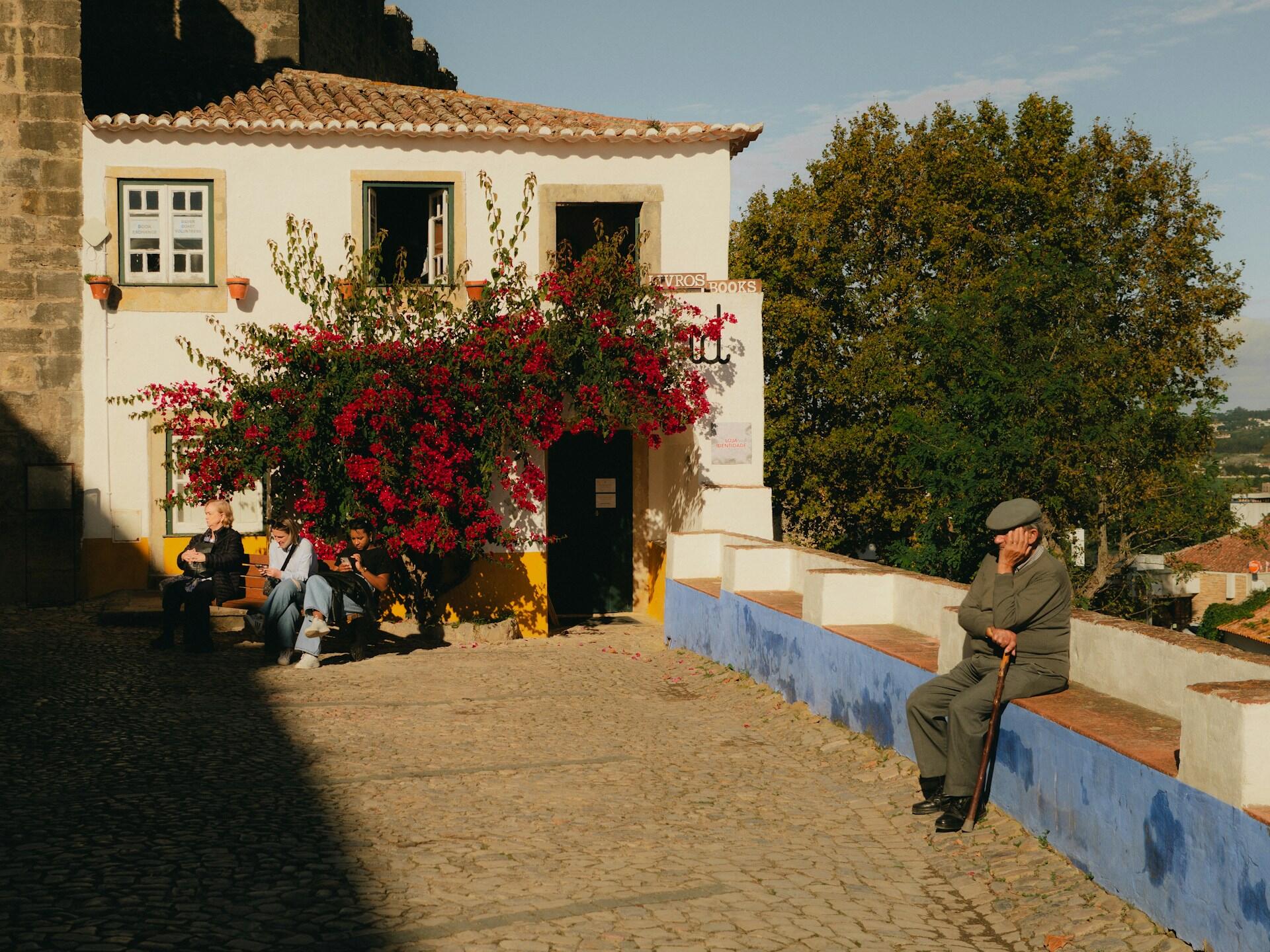 A quaint street scene features a charming white building adorned with vibrant bougainvillea, while people sit and relax on the cobblestones.
