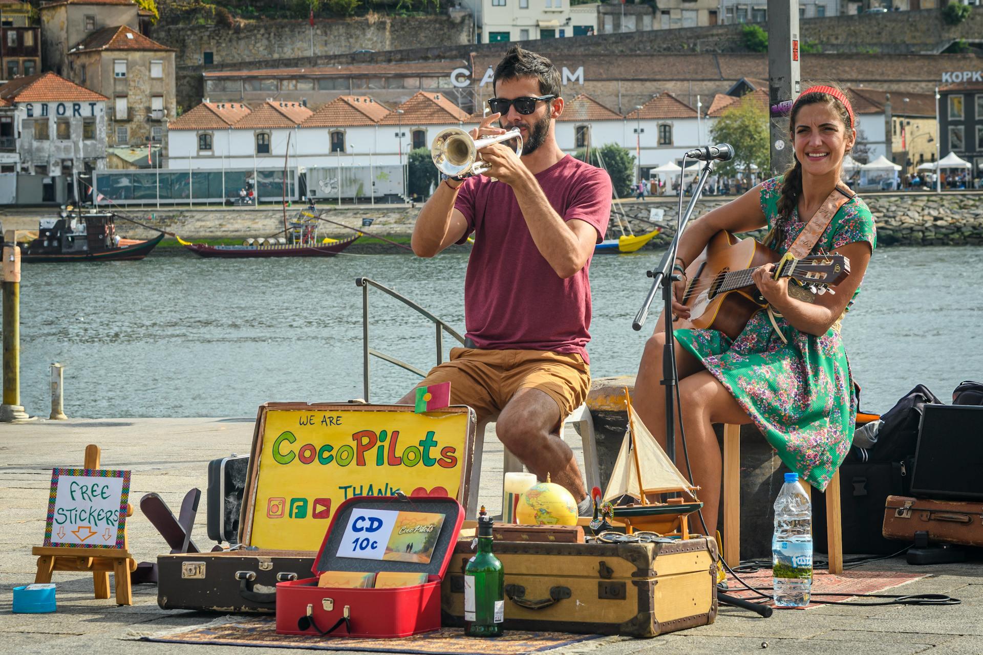 A duo of street performers play music by the riverside, with a colorful sign promoting their band and merchandise.