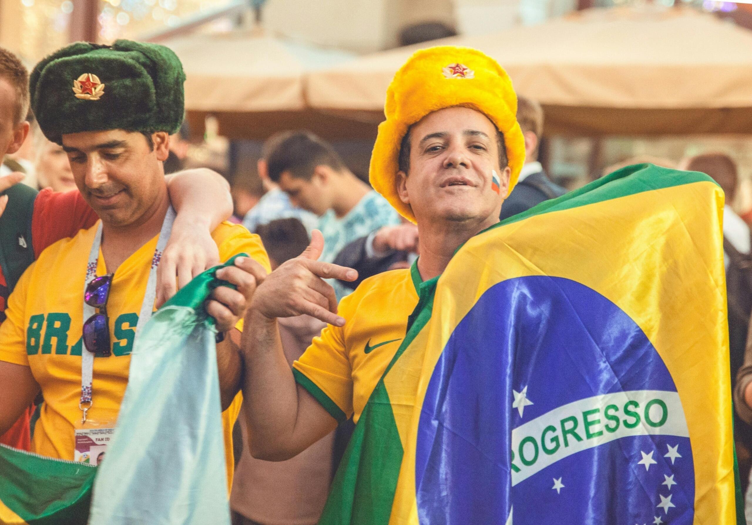 Two men wear Brazil soccer fan clothing. One holds the flag of Brazil.
