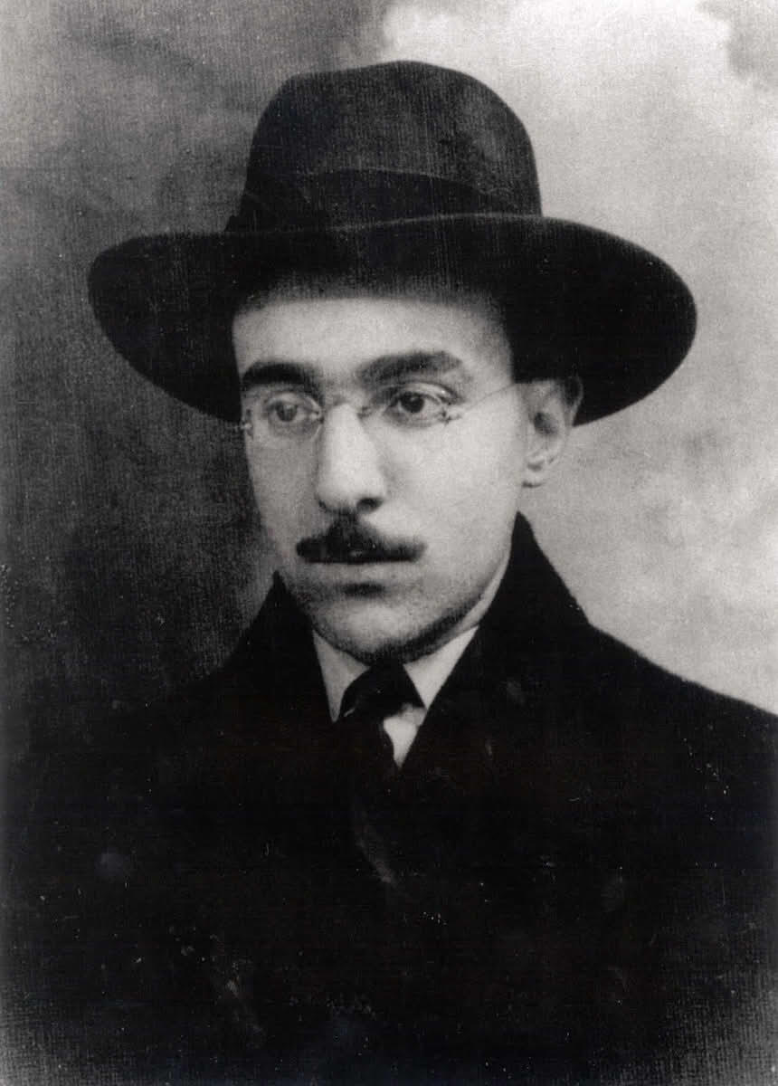 A black-and-white portrait of a man wearing a wide-brimmed hat, round eyeglasses, a mustache, and a dark overcoat with a tie. He has a serious expression and appears to be from the early 20th century.