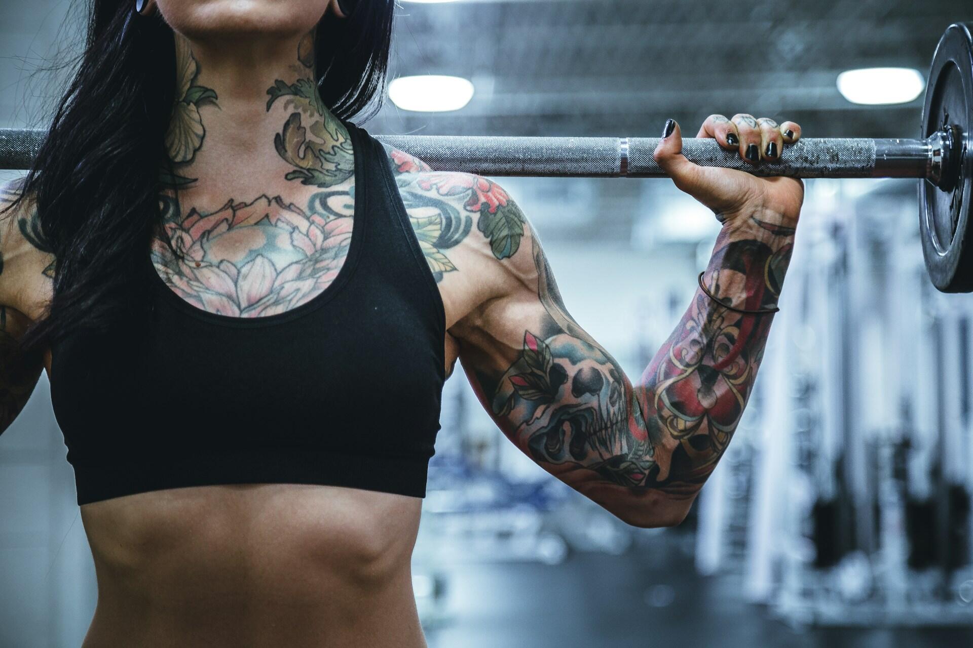 A muscular person with colorful tattoos holds a barbell at shoulder height in a gym setting, showcasing strength and dedication.