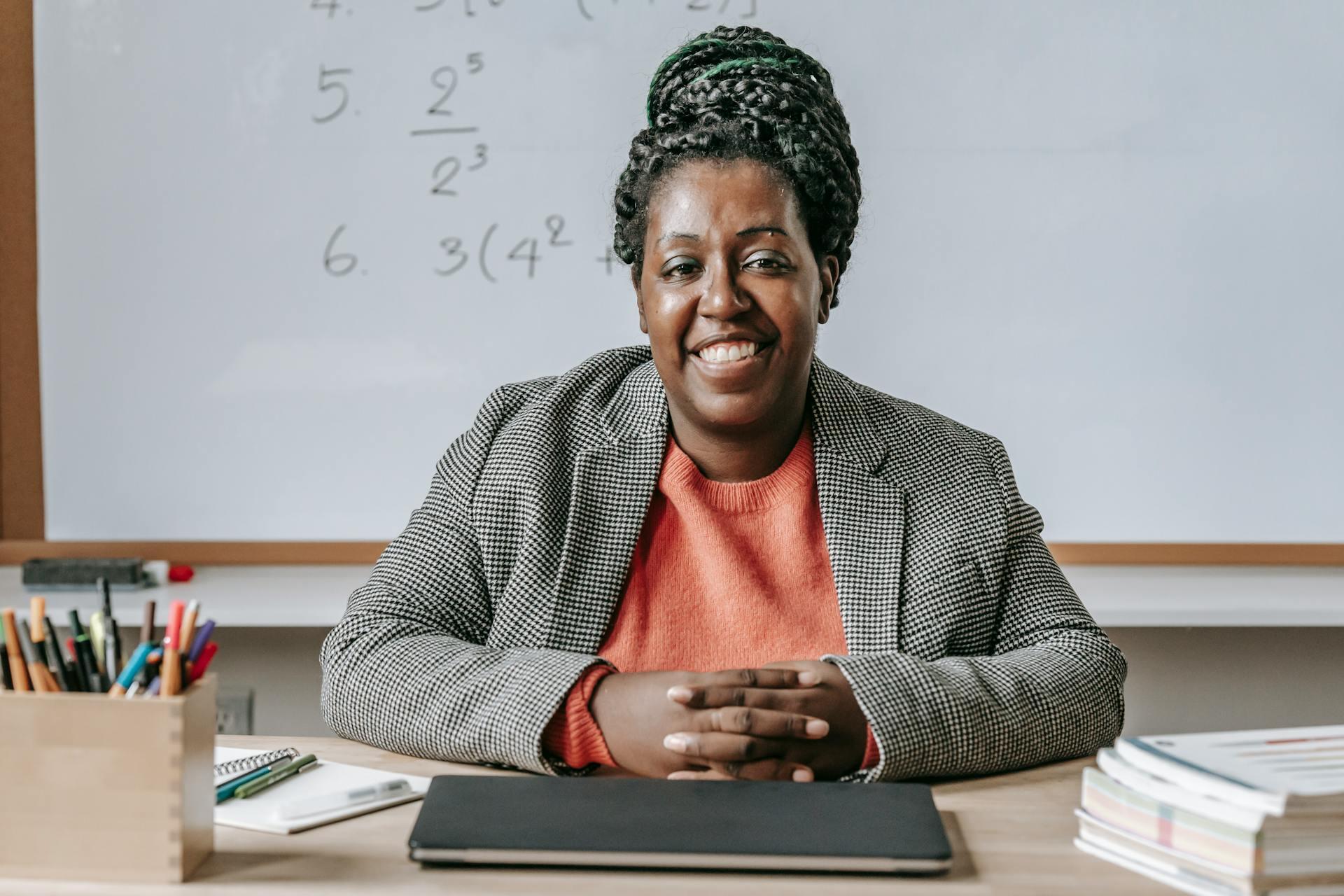 Teacher with braided hair sits at desk with closed laptop, books, and pens, in front of whiteboard with math equations.