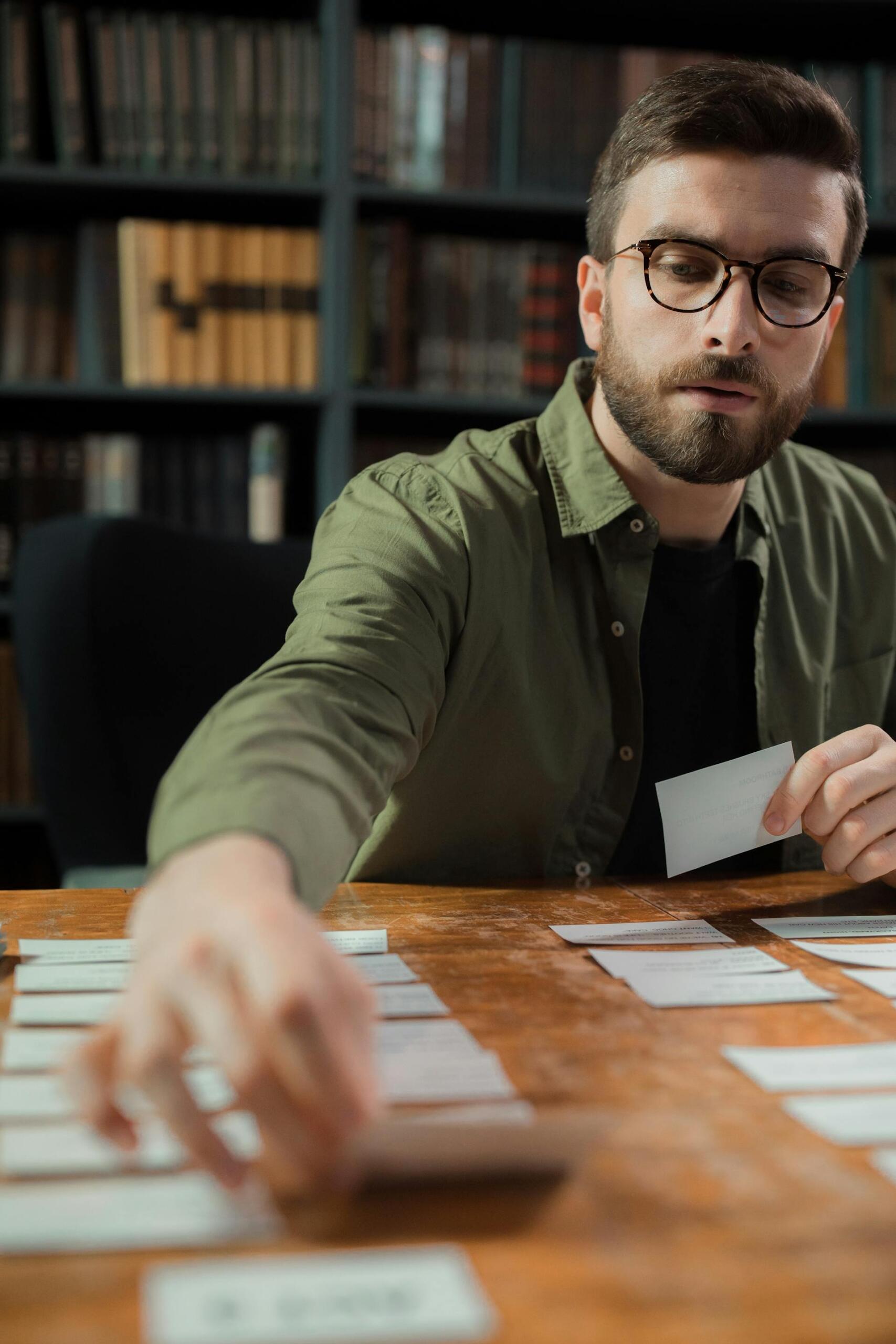 Person in a green shirt organizing index cards on a wooden table with bookshelf in the background.