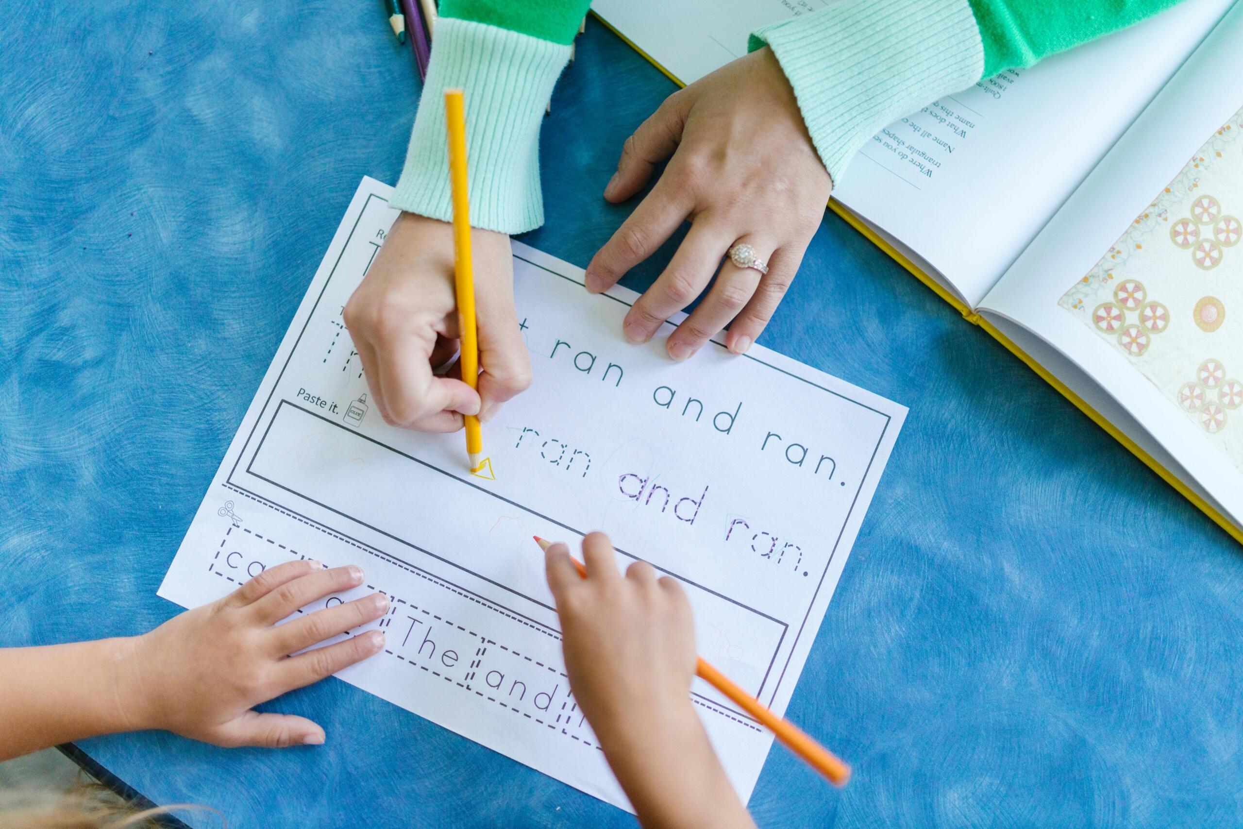 Close-up of a child and an adult working together on a reading worksheet, with the adult writing on the sheet and the child following along.