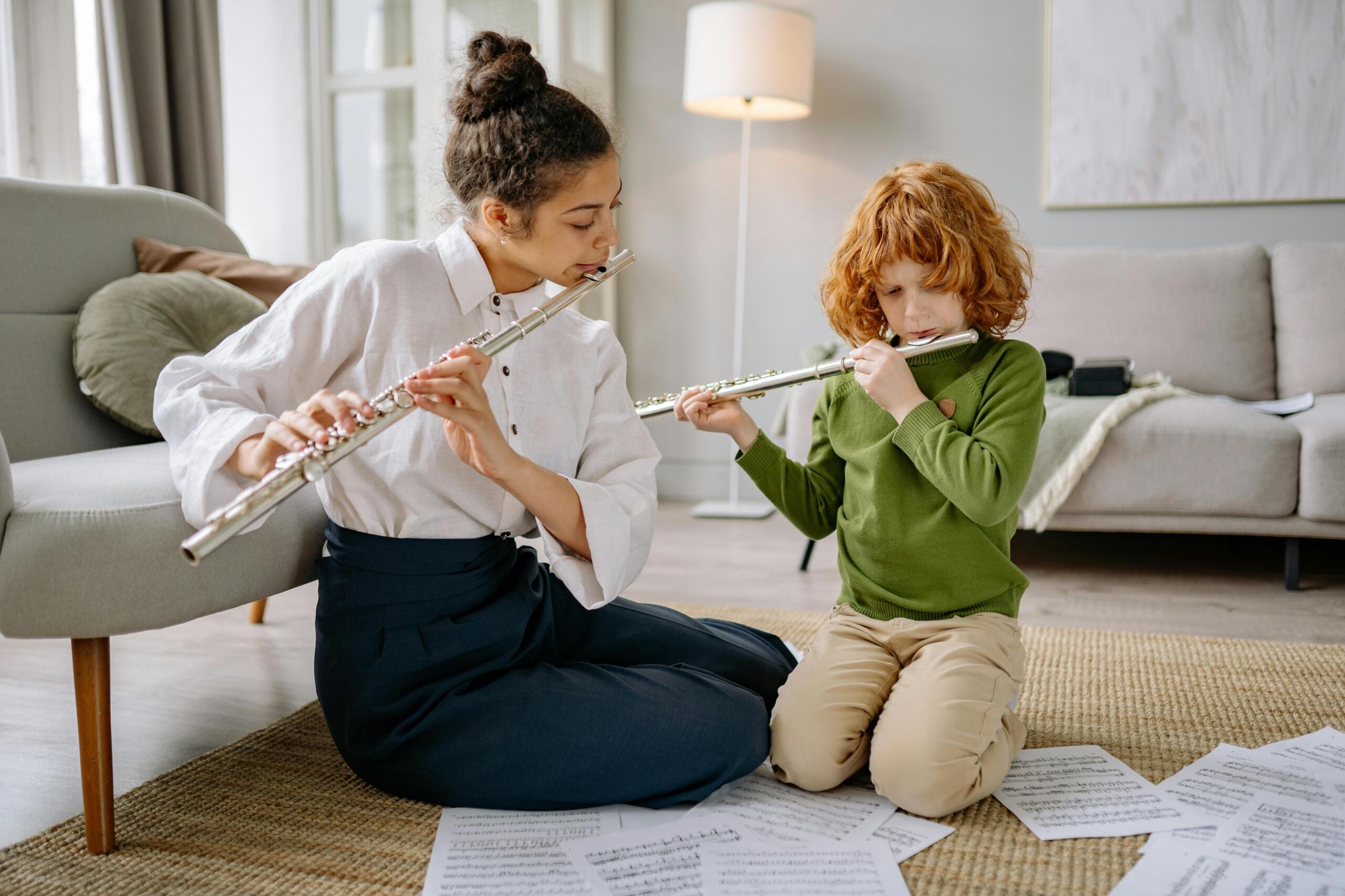 A woman teaching a young boy how to play the flute, both sitting cross-legged on the floor in a relaxed, home setting